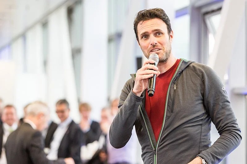 Ein junger Mann spricht in einem Mikrofon bei einem Event oder einer Konferenz, im Hintergrund sind mehrere Frauen und Männer in formelle Kleidung sichtbar.