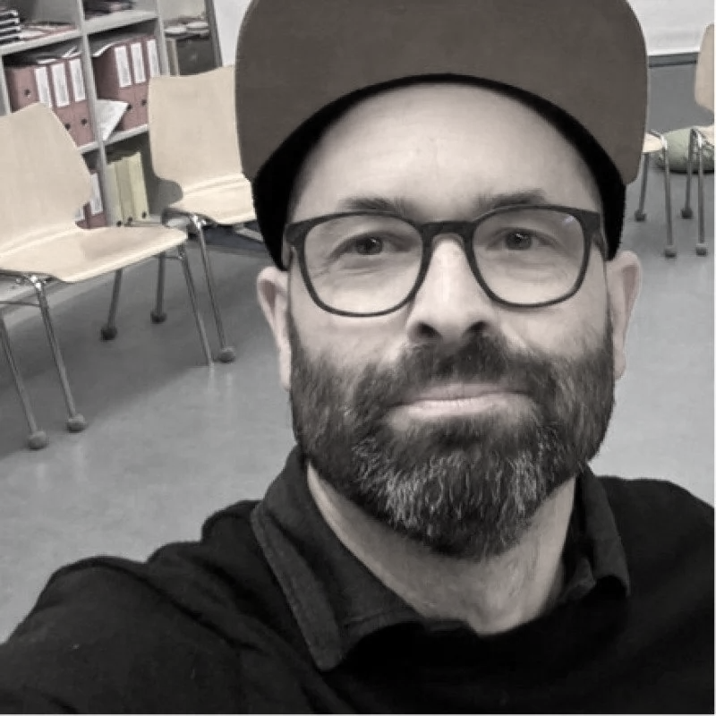 Ein Mann mit Brille und Bart, trägt eine Cap, in einem Raum mit grauem Boden, leeren Stühlen und einer Bücherregalwand im Hintergrund.