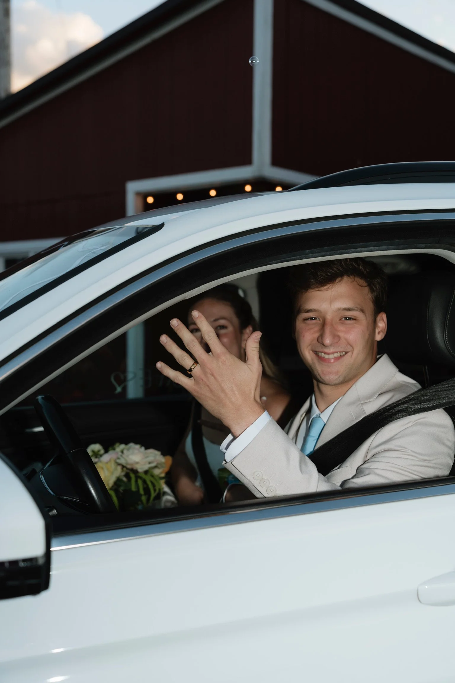 Young man in a light-colored suit smiling and waving from the driver's seat of a white car during a wedding, with a woman behind him and a building with a dark red exterior and string lights in the background.