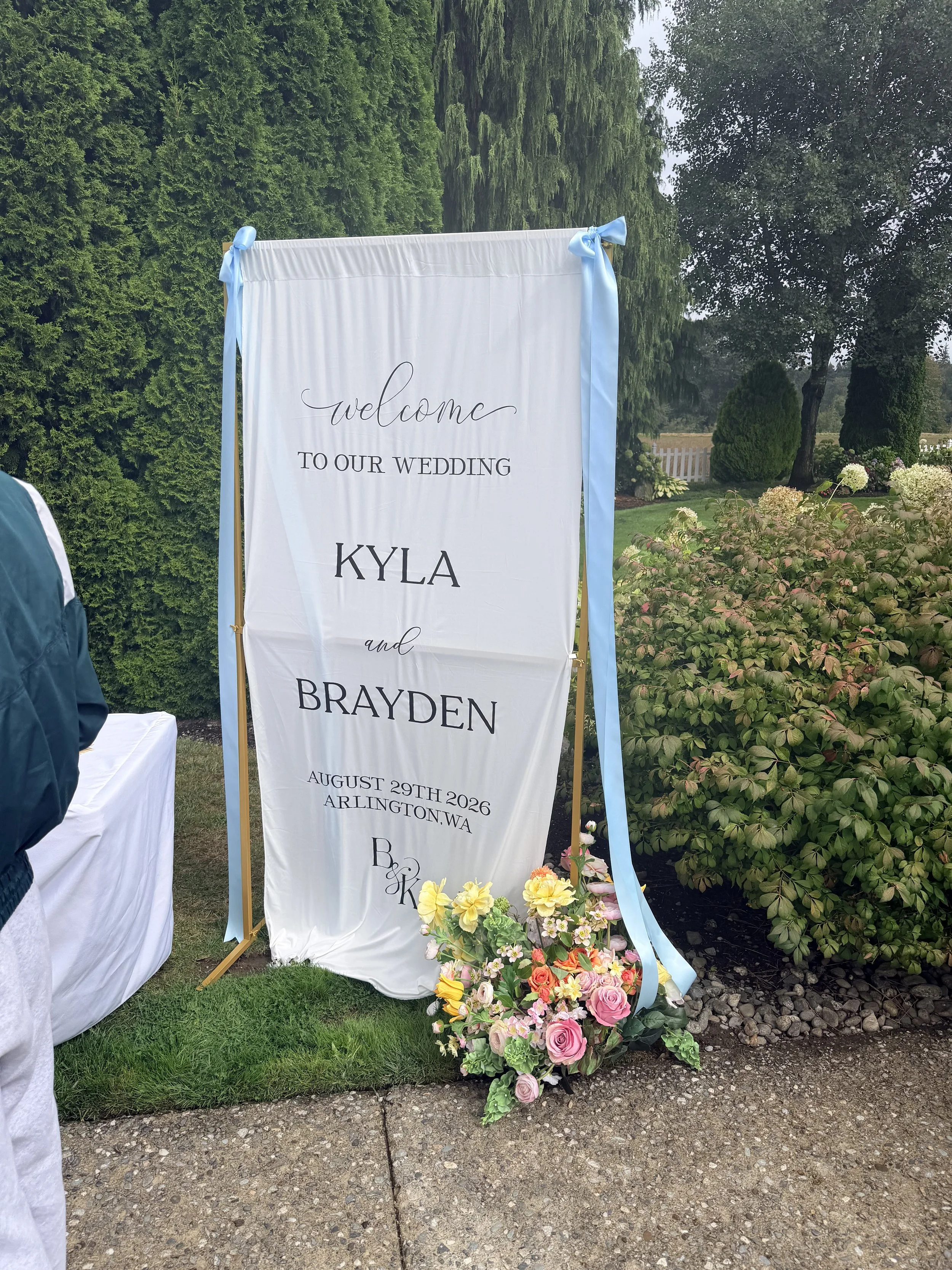 A wedding welcome sign with the names 'Kyla and Brayden' and the wedding date 'August 29th, 2026' in Arlington, Washington, displayed outdoors with flowers and greenery in the background.