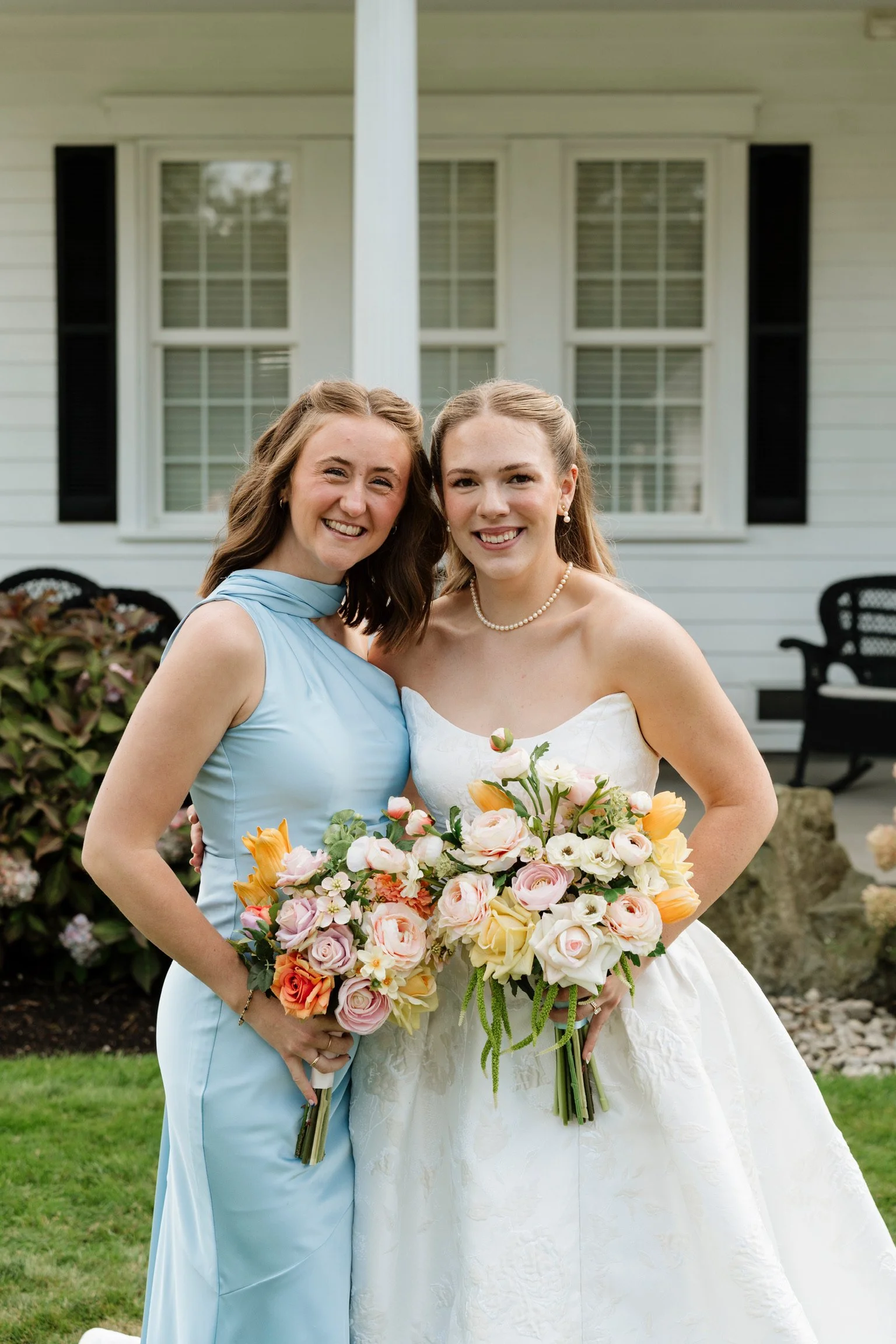 Two women are standing outside on grass in front of a house with white siding and windows. One is wearing a light blue dress with a matching scarf, and the other is wearing a white wedding dress with pearl earrings and necklace. They are smiling and 
