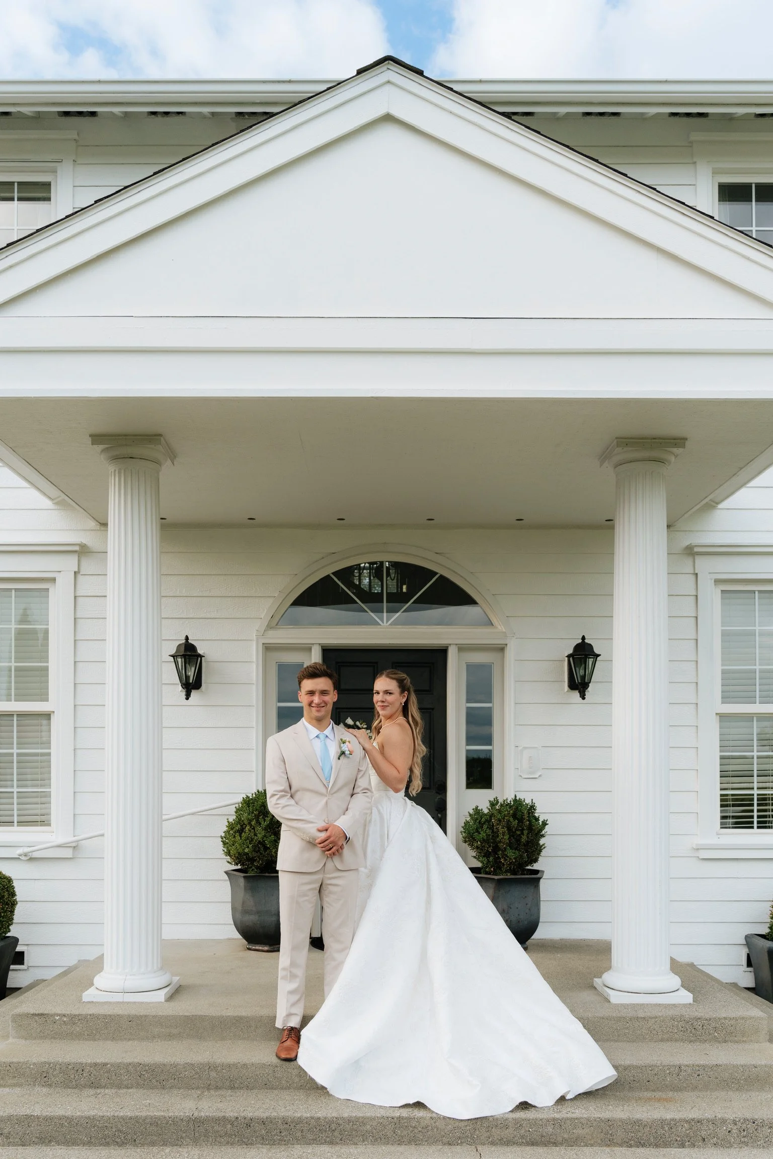 A smiling man in a beige suit and tie with a boutonniere standing next to a woman in a white wedding dress with a long train, both standing on the steps of a white house with columns and black lantern lights.