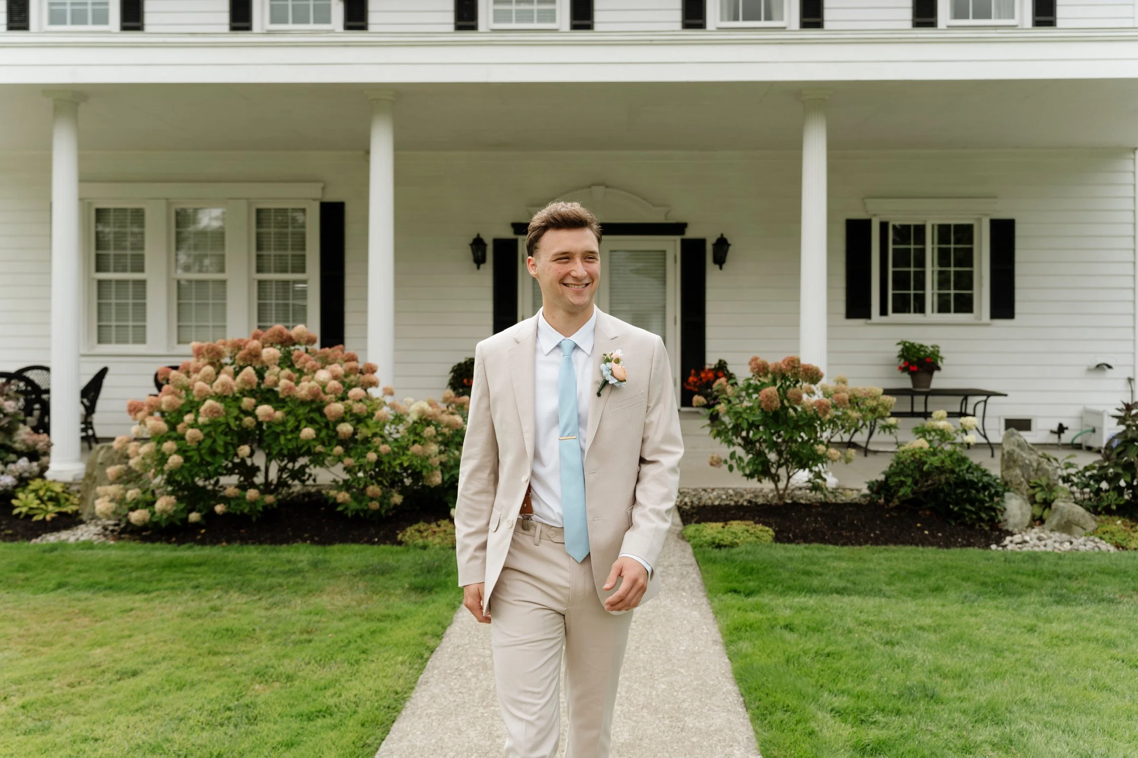 A man in a beige suit with a light blue tie and boutonniere smiling and walking outside in front of a white house with black shutters, surrounded by bushes and flowers.