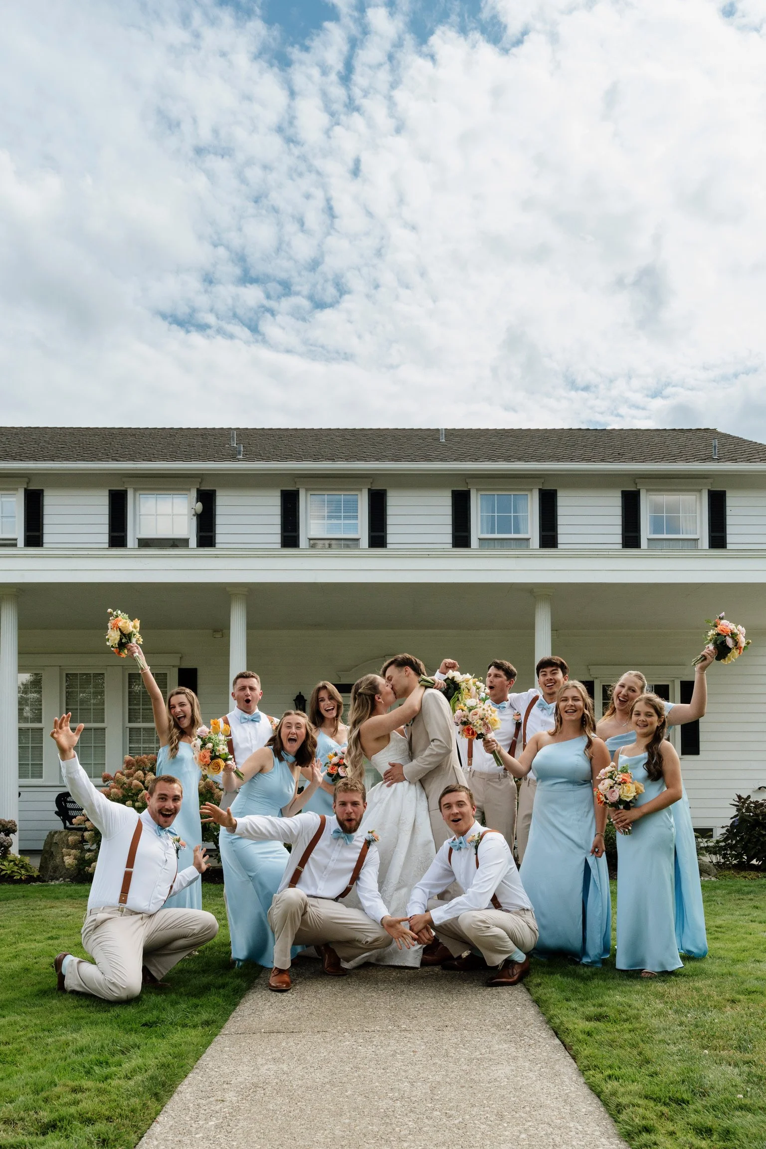 A group of people celebrating a wedding outside a house, with a man and woman kissing in the center, surrounded by friends holding bouquets and smiling.