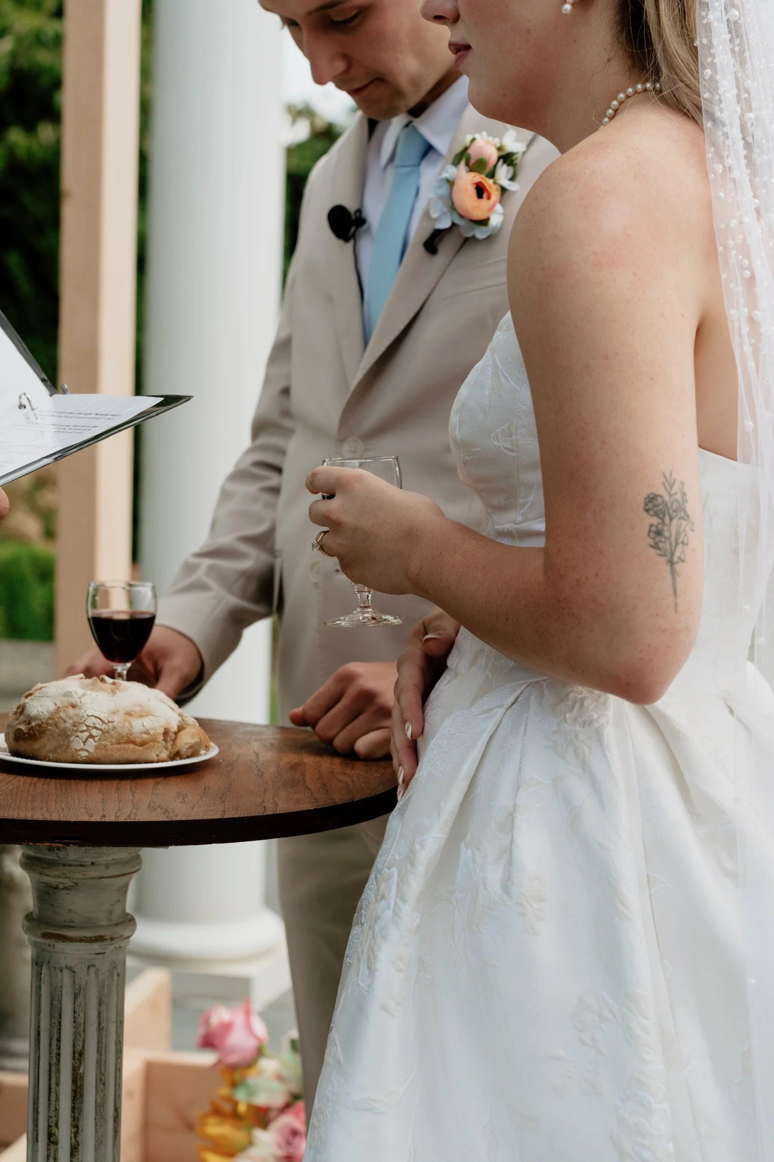 A bride and groom standing together at their wedding ceremony, holding glasses of wine, with a table of bread and a officiant reading from a paper, outdoors with greenery and flowers in the background.