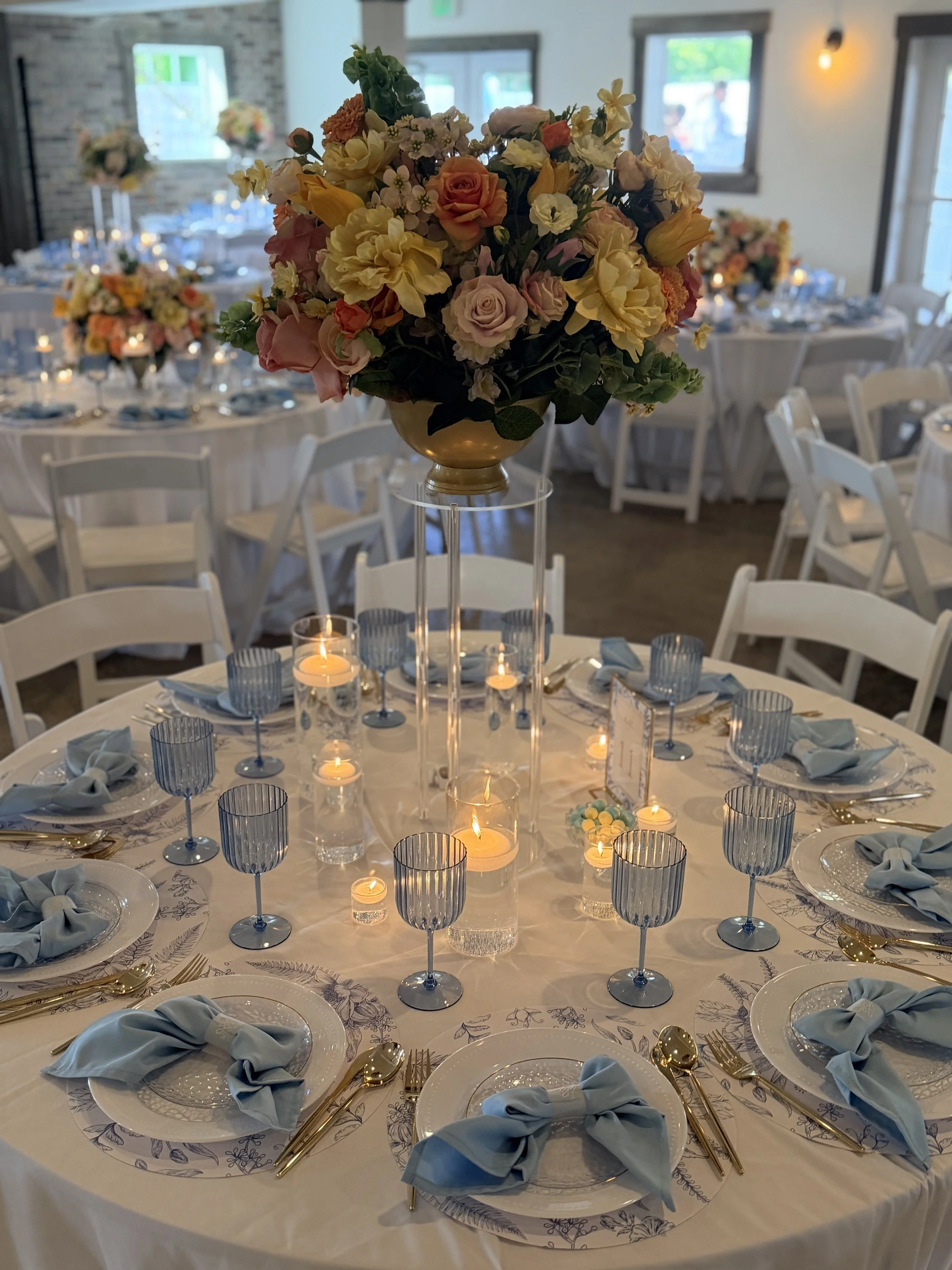 Elegant banquet table decorated with a floral centerpiece, candles, blue glassware, and place settings with napkins tied in bows, set in a well-lit room with windows and additional floral arrangements in the background.