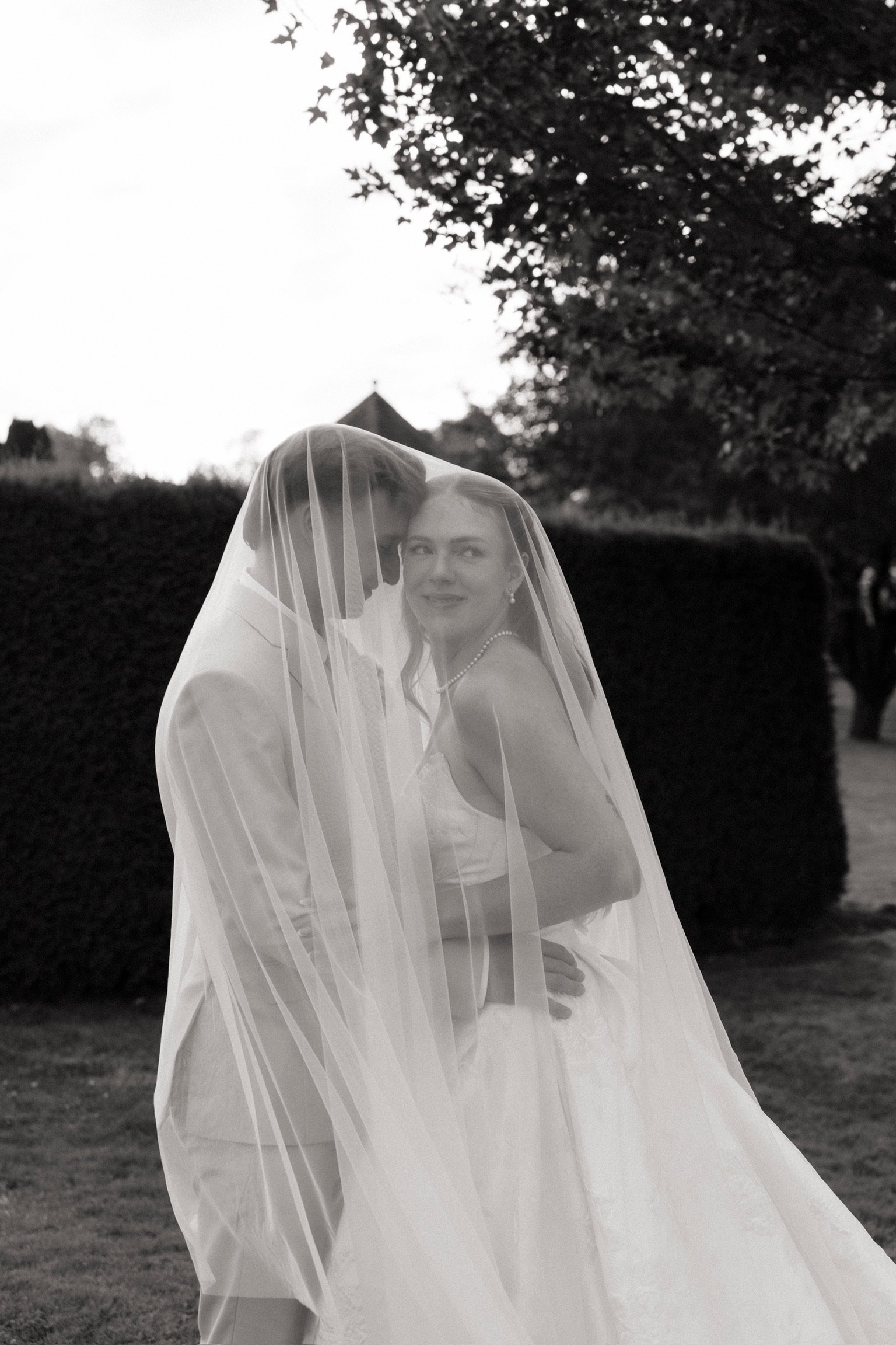 A bride and groom sharing a private moment outdoors, with the groom kissing the bride on the forehead under her veil, and the bride smiling gently, surrounded by trees and bushes in the background.