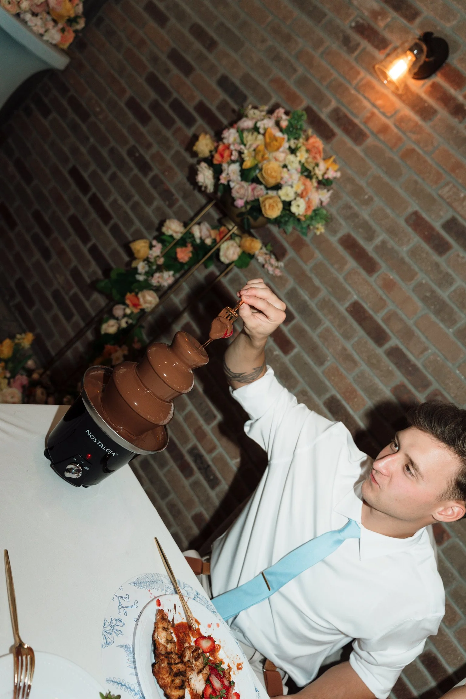 A man in a white shirt and light blue tie is sitting at a table with a plate of food and a chocolate fountain. He is dipping food into the fountain, which is in a chocolate warmer. The background shows a brick wall, a floral arrangement, and a vintag