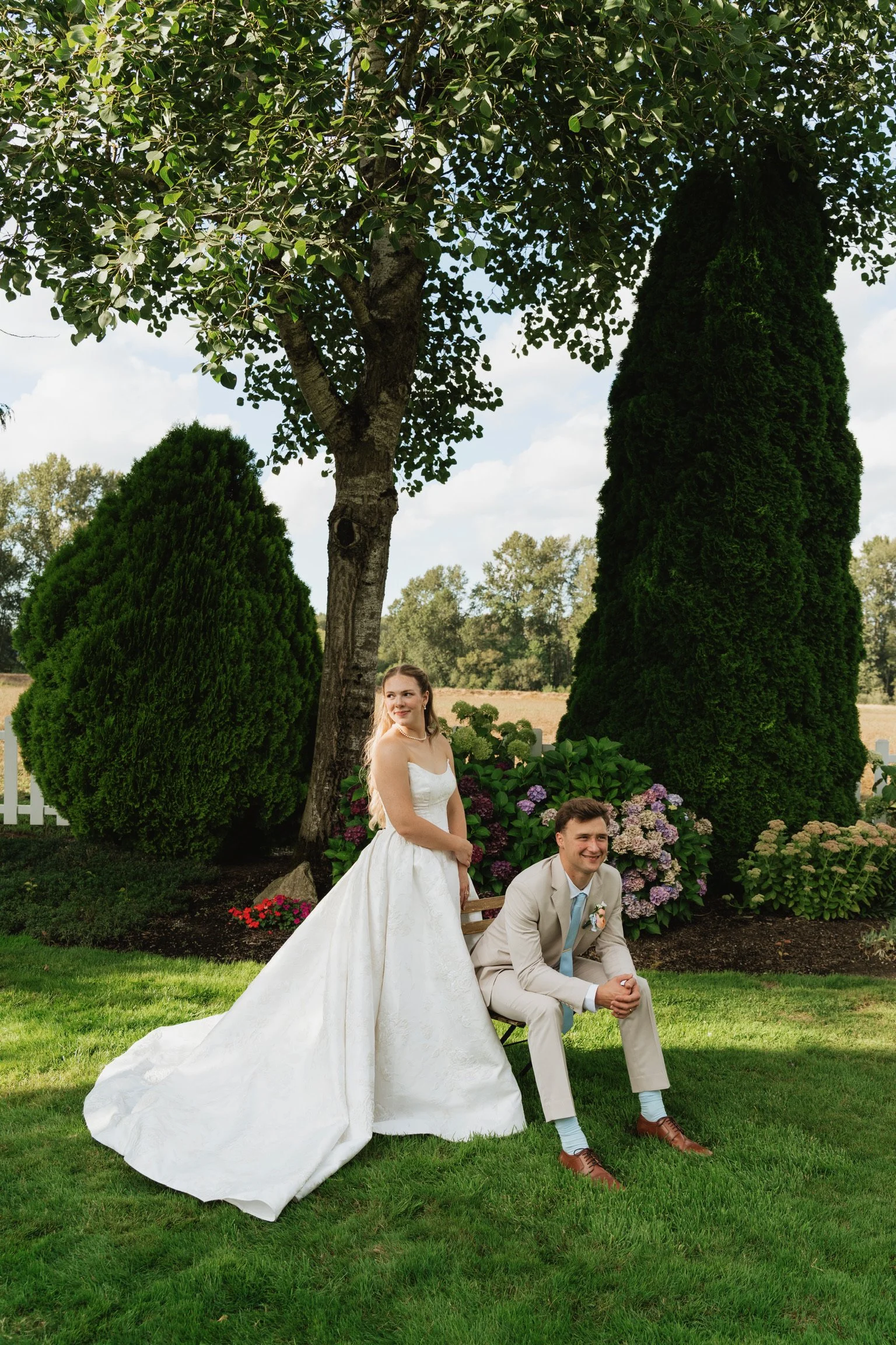 A bride in a white wedding gown and a groom in a beige suit with a blue tie are outdoors under a tree and bushes, posing for a photo. The bride is standing, looking away, and the groom is kneeling on the grass, smiling.