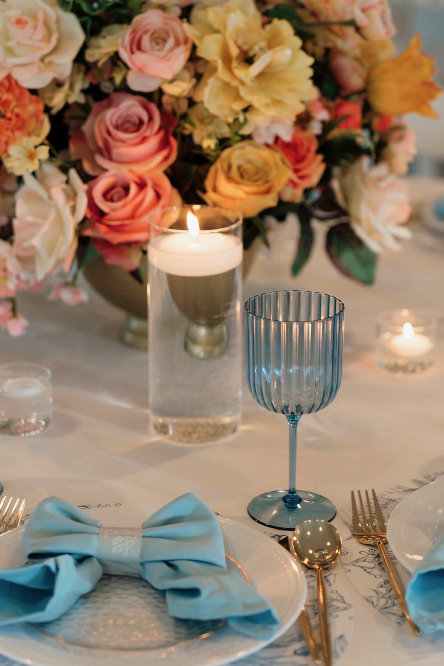 A table setting with a large floral centerpiece of pink, yellow, and white roses and other flowers. There are candles, including a tall white one and smaller teal-colored ones. The table has a blue napkin folded on a white plate, gold cutlery, and a printed tablecloth.