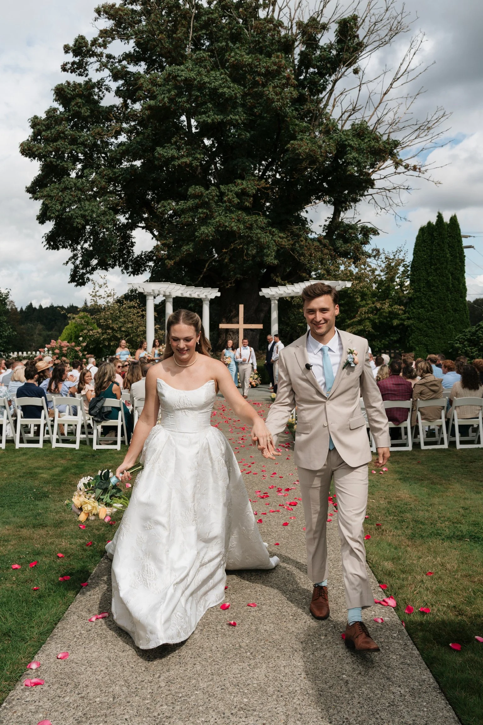 A newlywed couple walking down an outdoor aisle, holding hands, surrounded by wedding guests seated on white chairs, with a large tree and a white cross in the background, during a wedding ceremony.