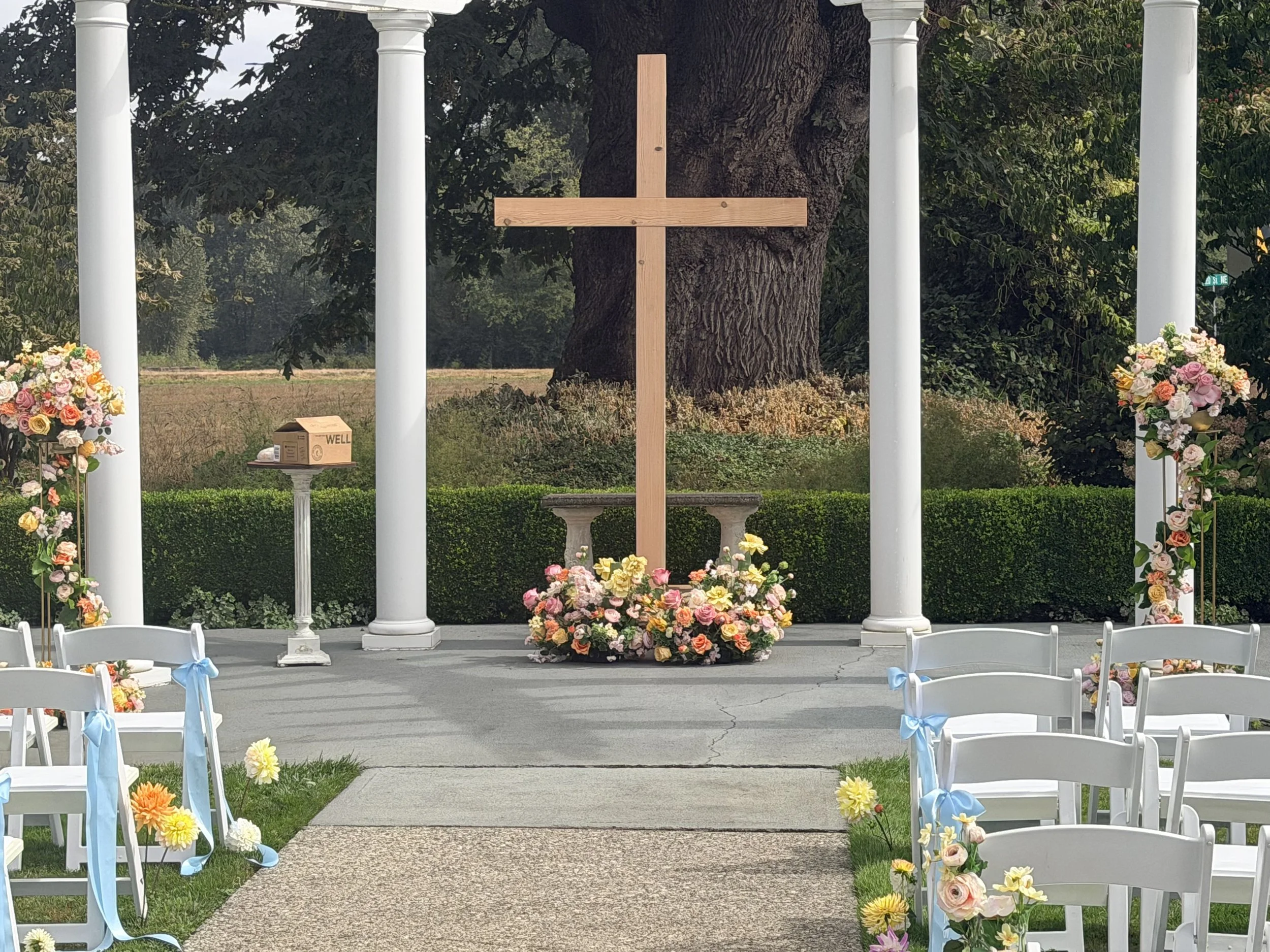 Outdoor wedding altar setup with a wooden cross in front of a large tree, decorated with colorful flower arrangements on white chairs and pillars.