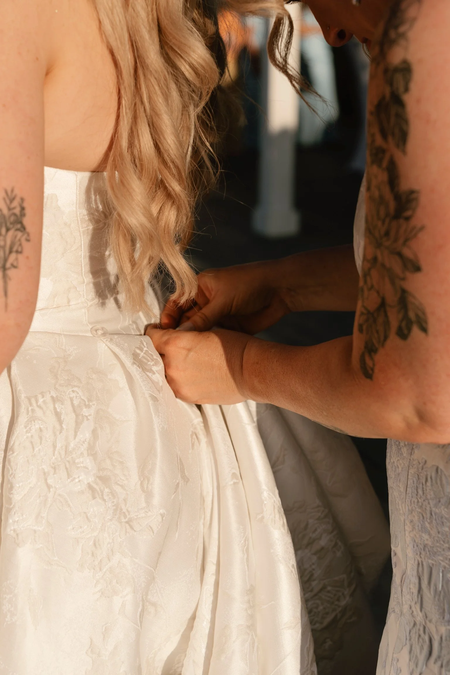 Person fastening a wedding dress on a bride, showing the back of the bride with blonde hair and tattoos on the arm.