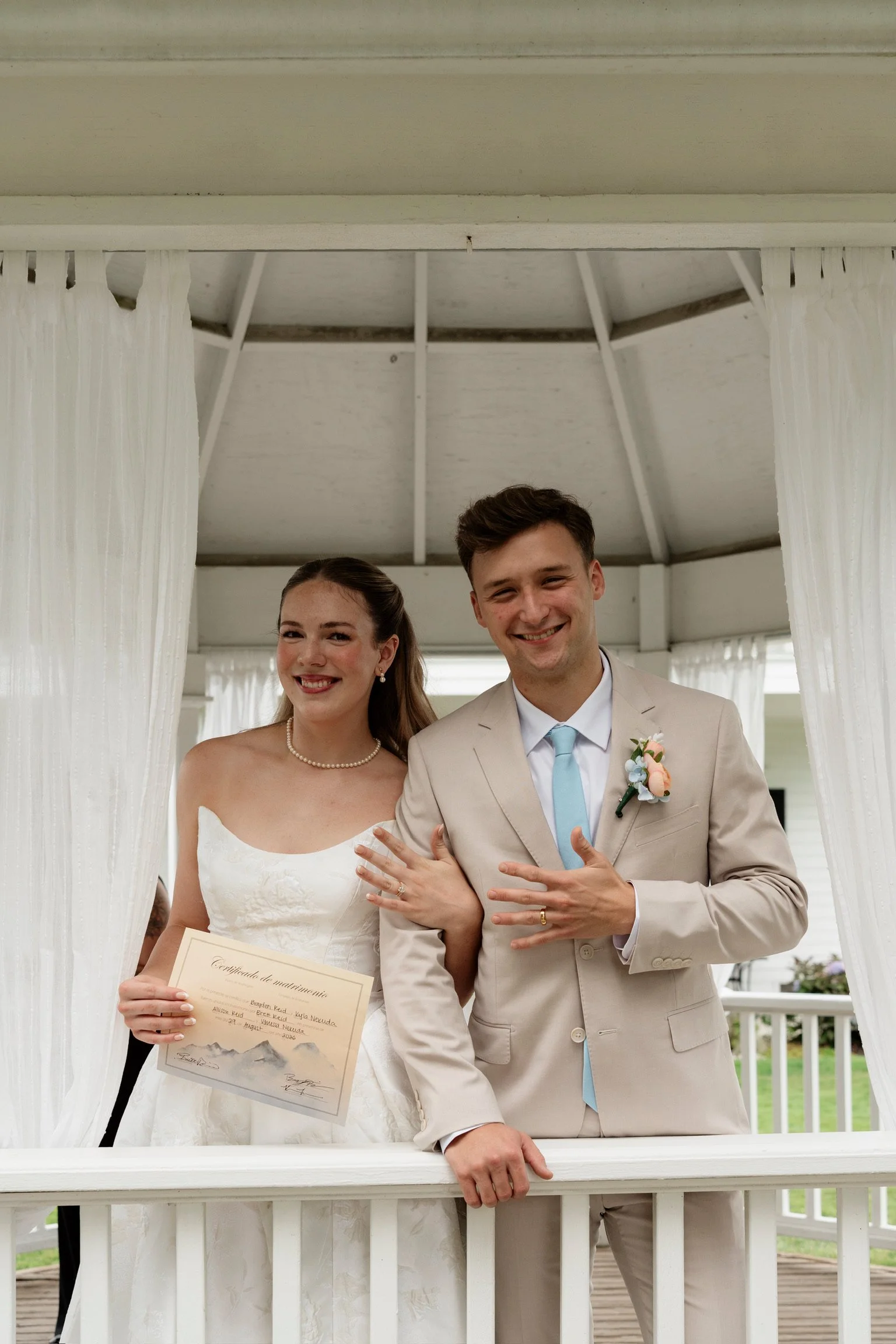 A bride and groom smiling and showing off their wedding rings during their wedding ceremony, standing behind a white railing with curtains and a roofed outdoor setting in the background.