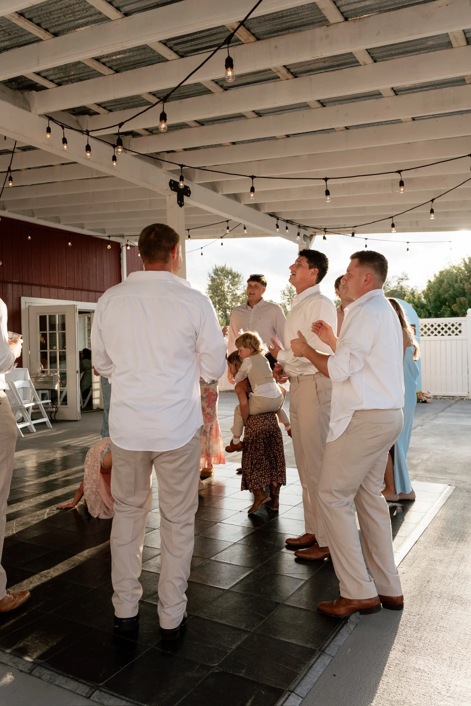 Group of people in white shirts and khaki pants dancing and socializing under a covered outdoor patio with string lights