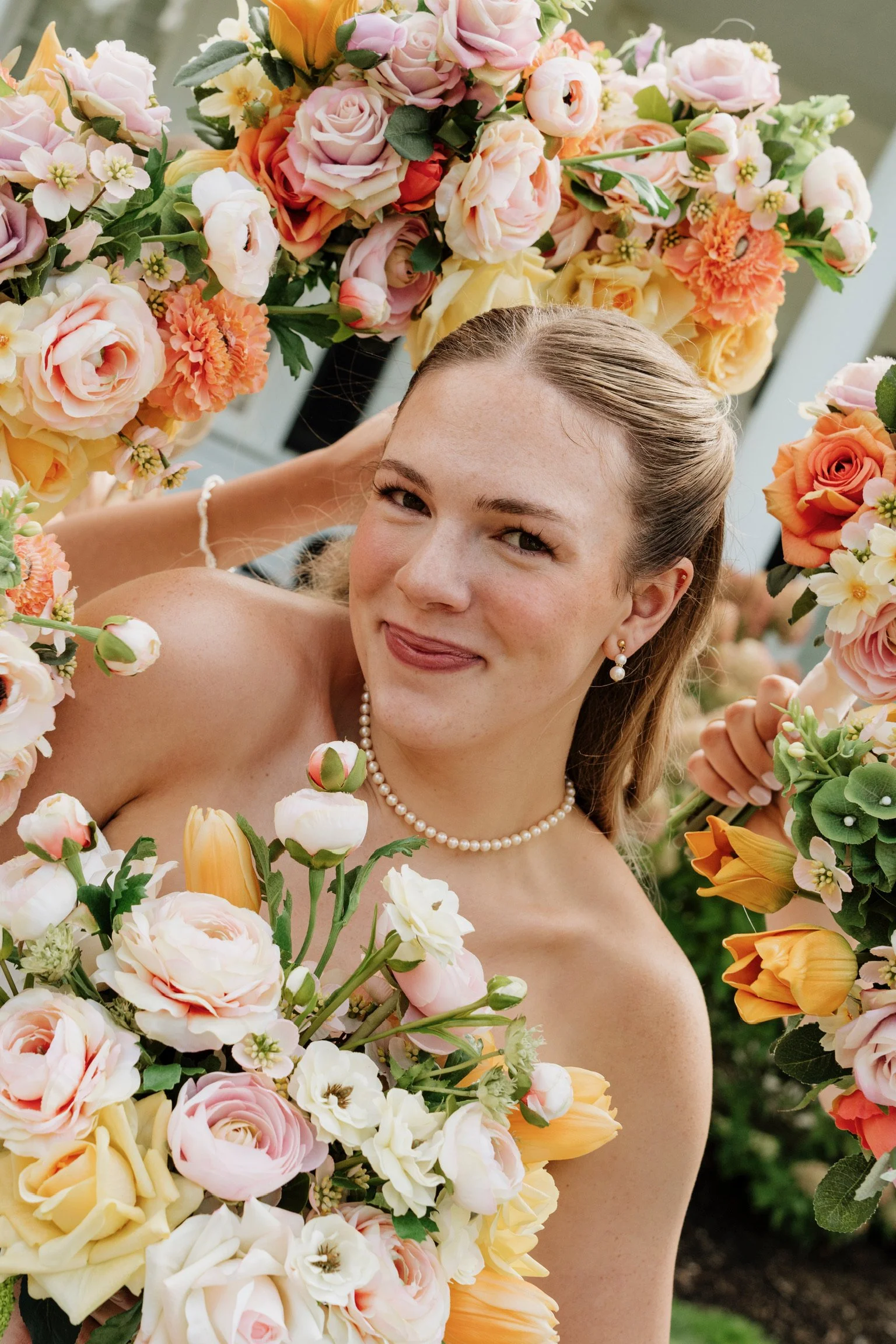A woman with blonde hair styled back, wearing pearl earrings and necklace, is smiling among a large arrangement of peach, pink, cream, and orange flowers, including roses, lilies, and other blossoms, outdoors.