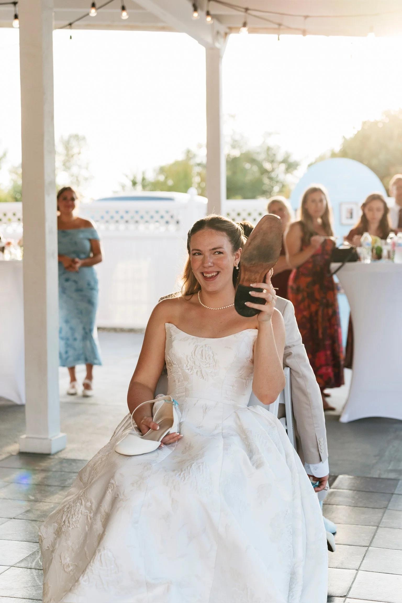 A smiling bride in a wedding dress sitting and holding a pair of white shoes, with a champagne bottle in her lap, at an outdoor reception with other guests in the background.