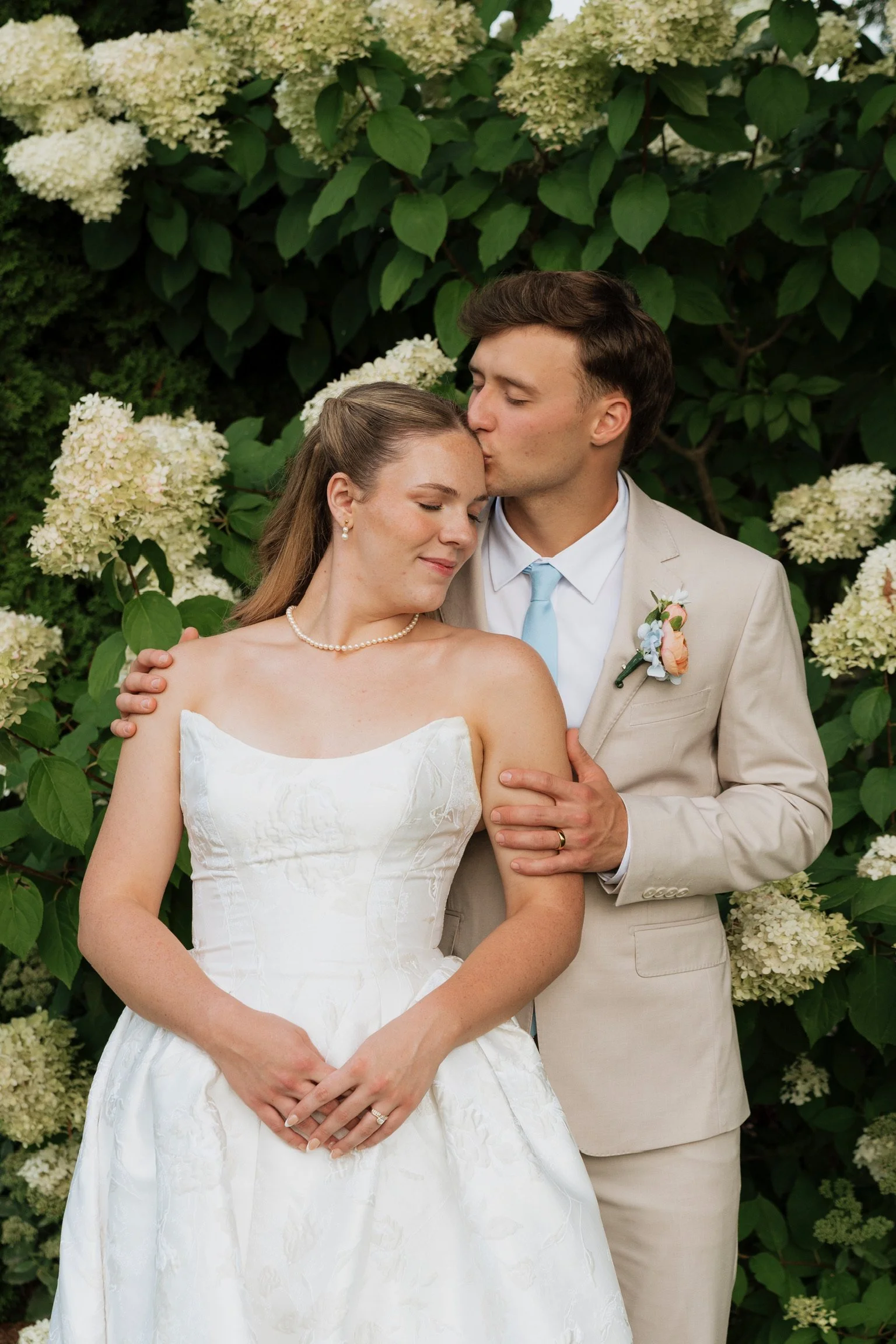 A newlywed couple standing together outdoors, surrounded by white hydrangea flowers and green leaves. The groom is kissing the bride on her forehead, and they both have their eyes closed, smiling softly. The bride is wearing a white strapless wedding