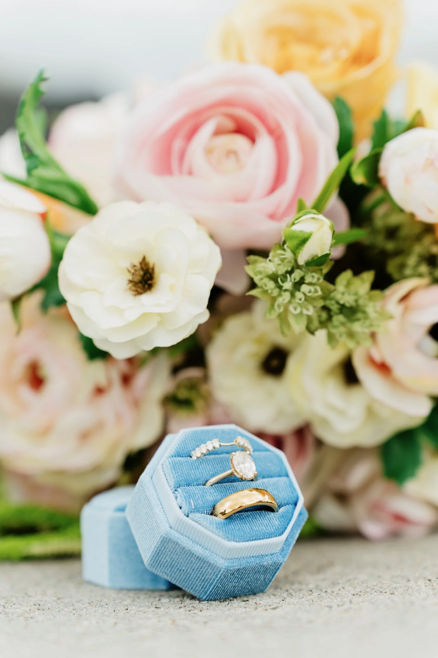A display of three rings on a blue velvet ring holder in front of a bouquet of pink, white, and yellow flowers, with a blurred background.