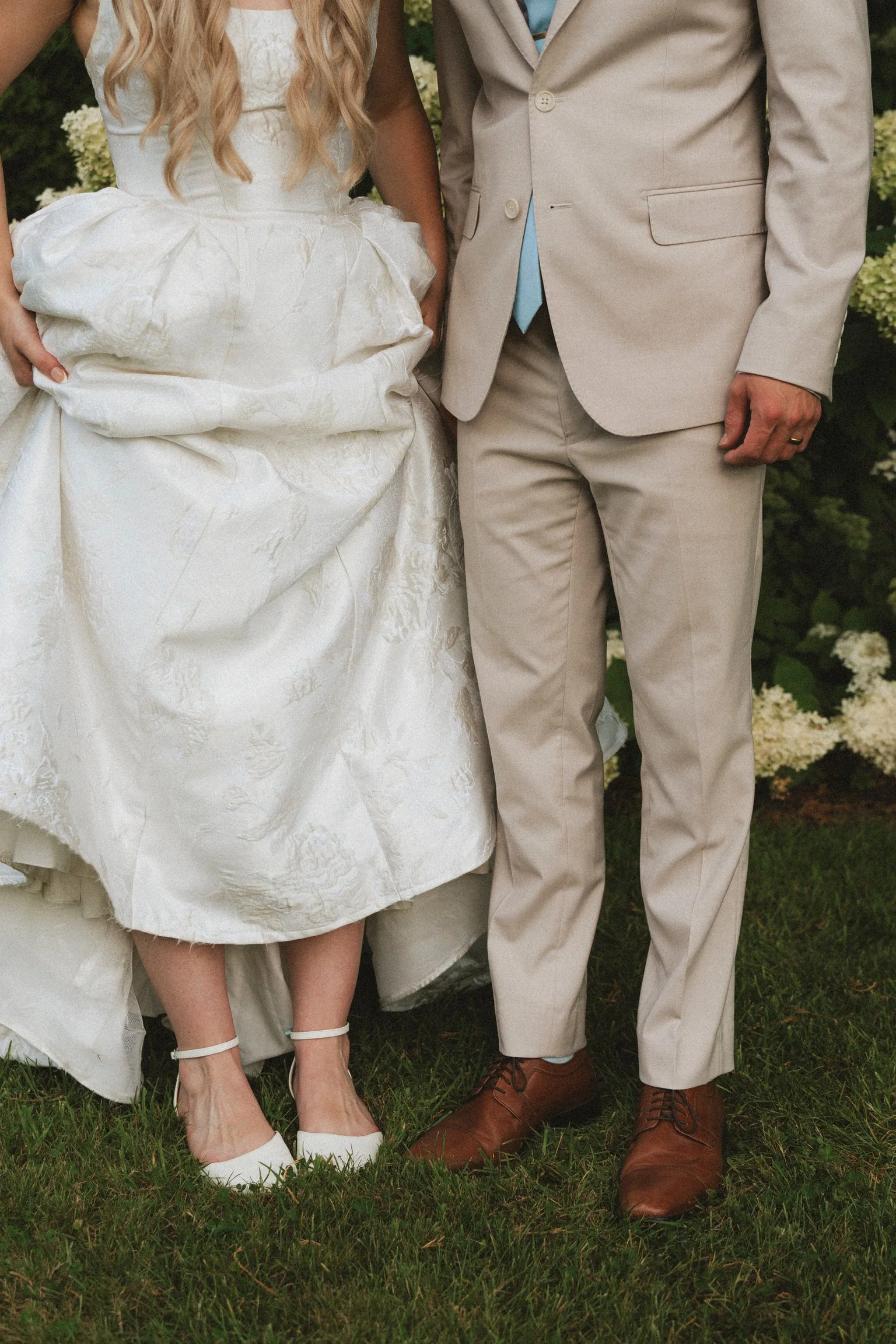Partial view of a couple dressed in wedding attire standing on grass; woman in a white wedding dress with heels, man in a beige suit with brown shoes.