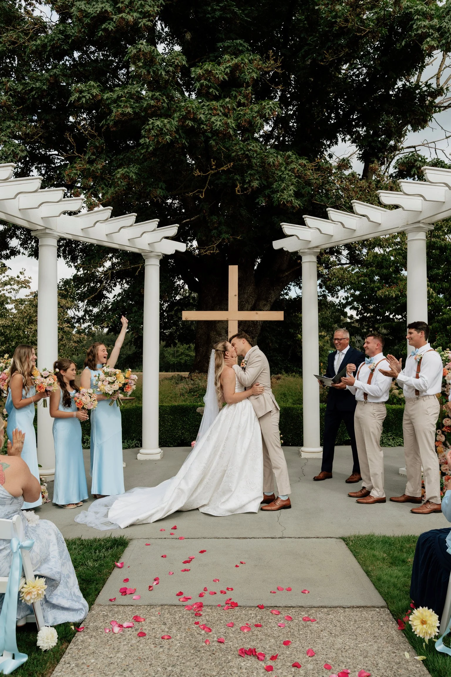 A wedding ceremony outdoors with a bride and groom kissing under a wooden cross, surrounded by bridesmaids holding bouquets and groomsmen clapping, with rose petals on the ground, a large tree in the background, and a white pergola overhead.