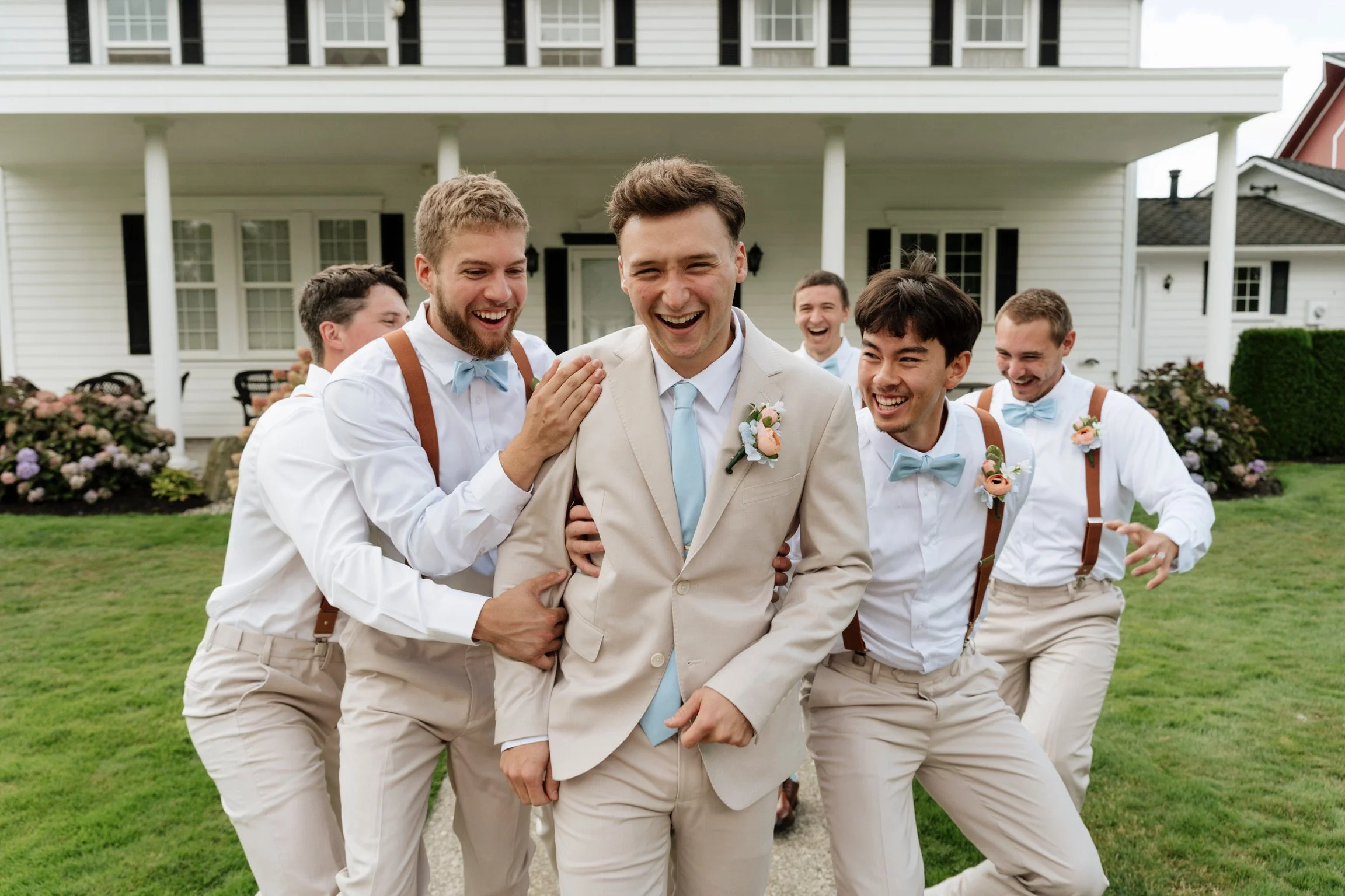 A group of men celebrating outdoors in front of a white house, with some wearing suspenders and bow ties, and one in a beige suit with a boutonniere, smiling and laughing.