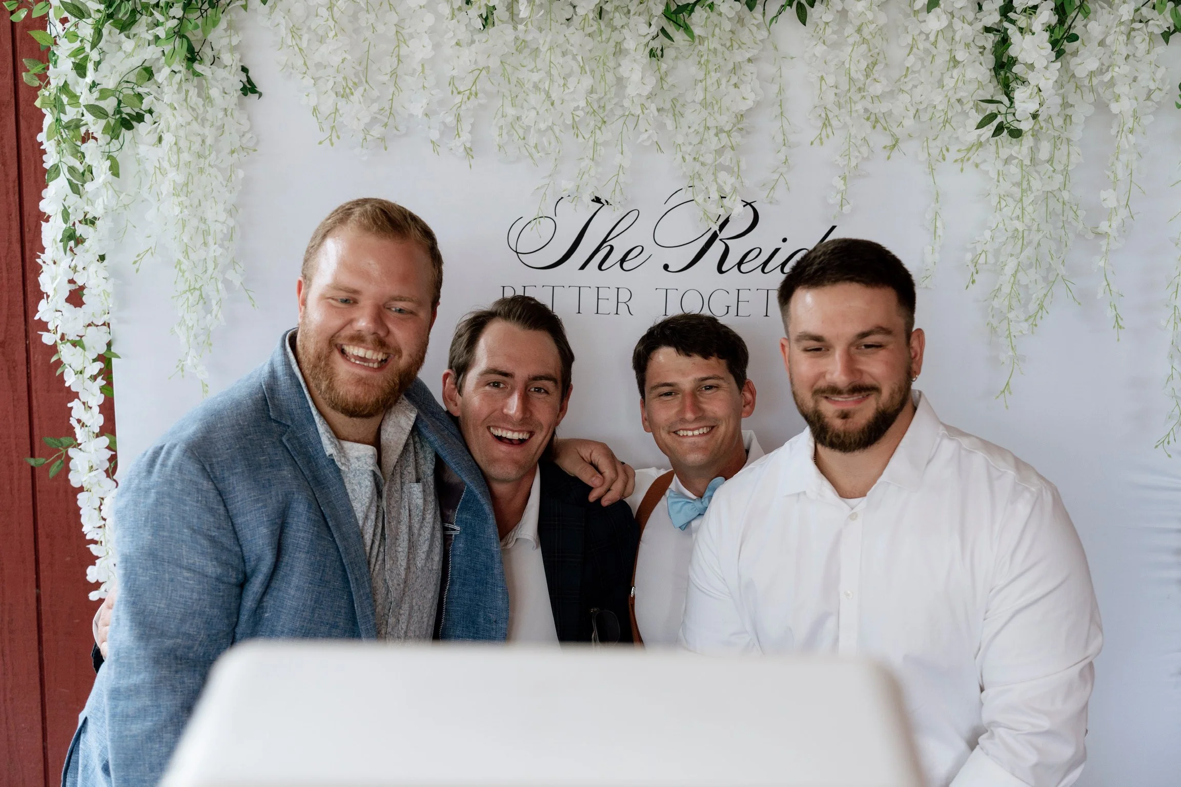 Four men smiling and taking a selfie in front of a floral backdrop with the text 'The Reids Better Together.'