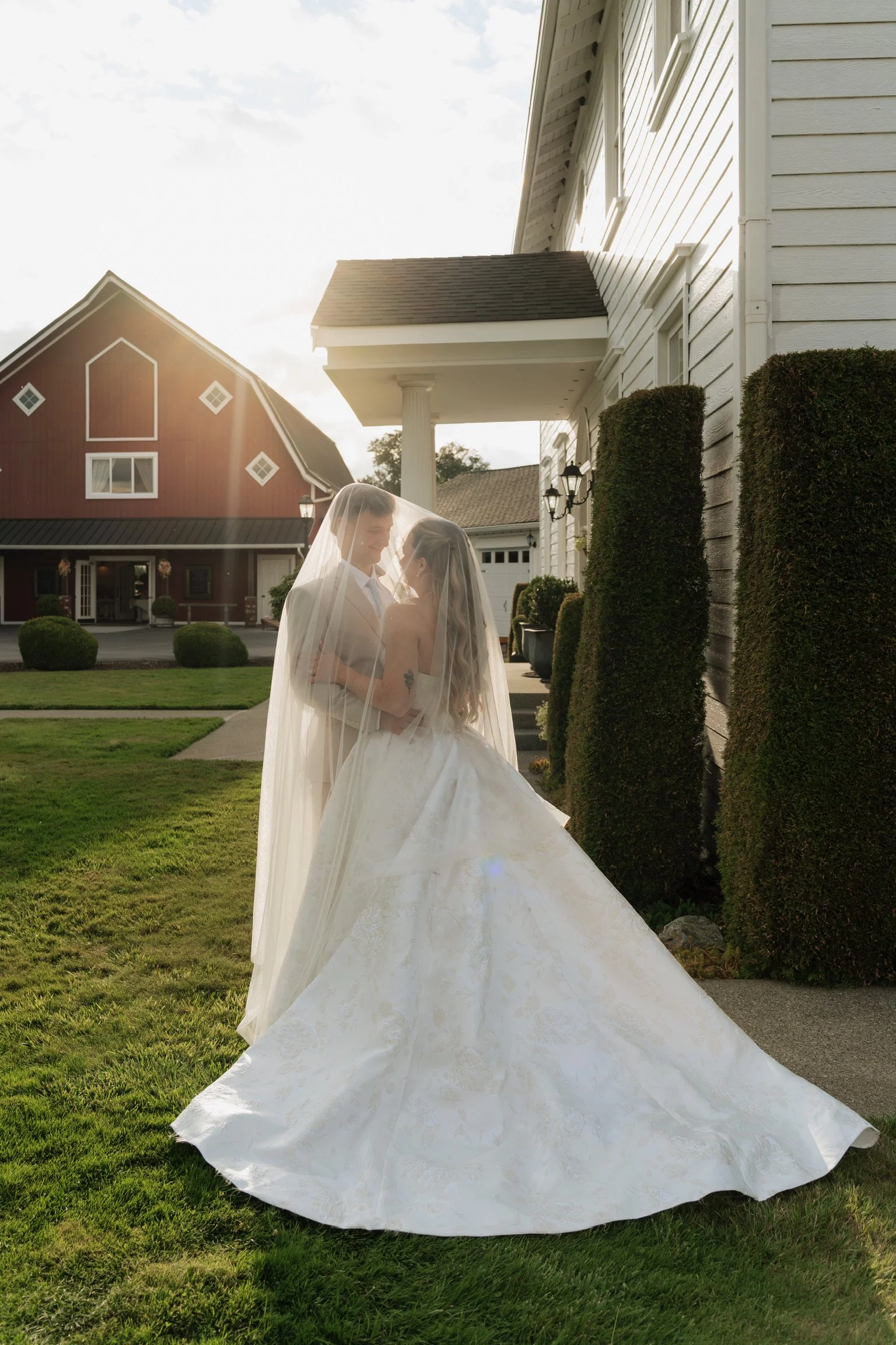 A bride and groom in wedding attire sharing a moment under a bridal veil outside a white house with greenery and a red barn in the background.