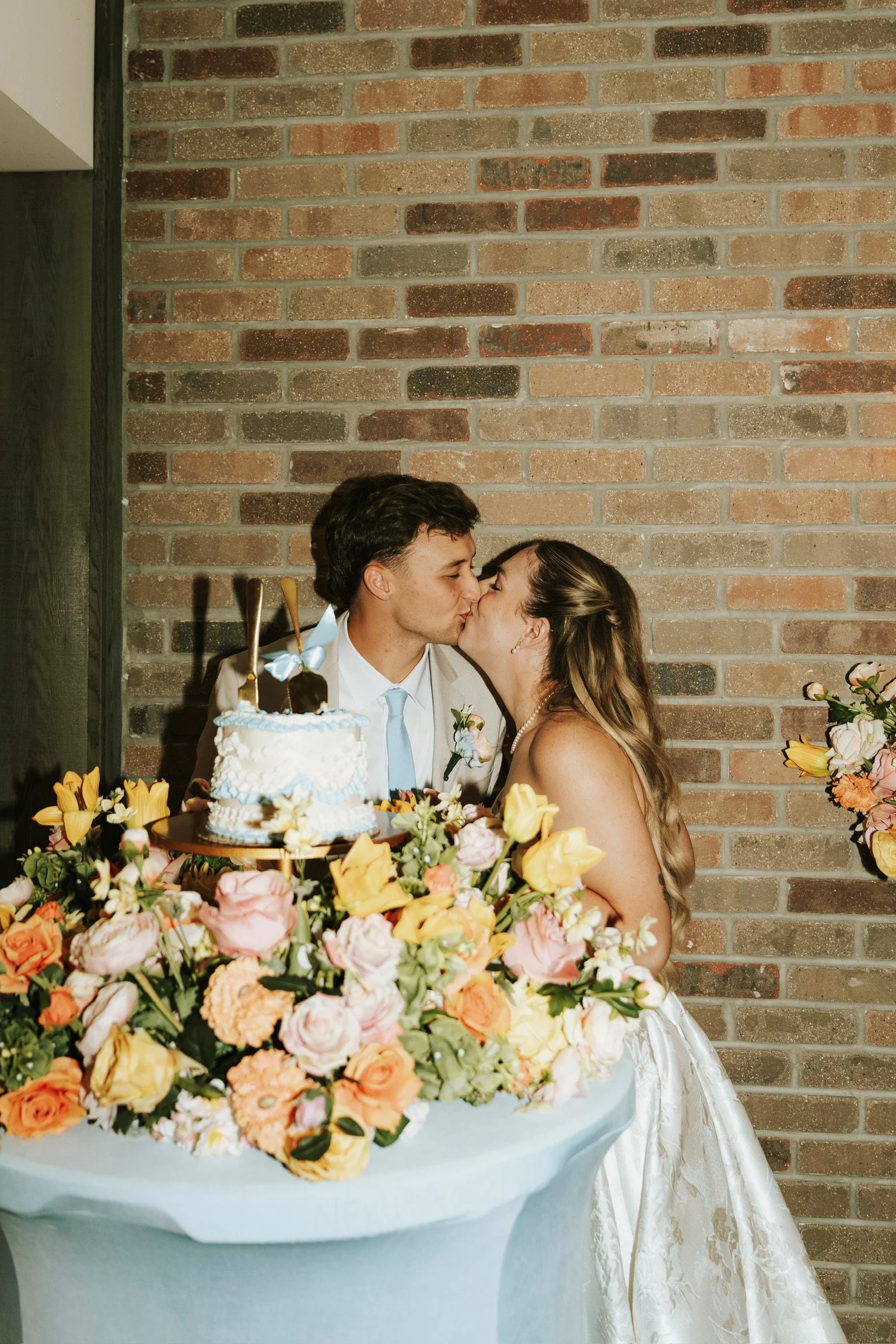 A bride and groom kissing in front of a brick wall during a wedding celebration, with a floral arrangement and wedding cake on the table in front of them.