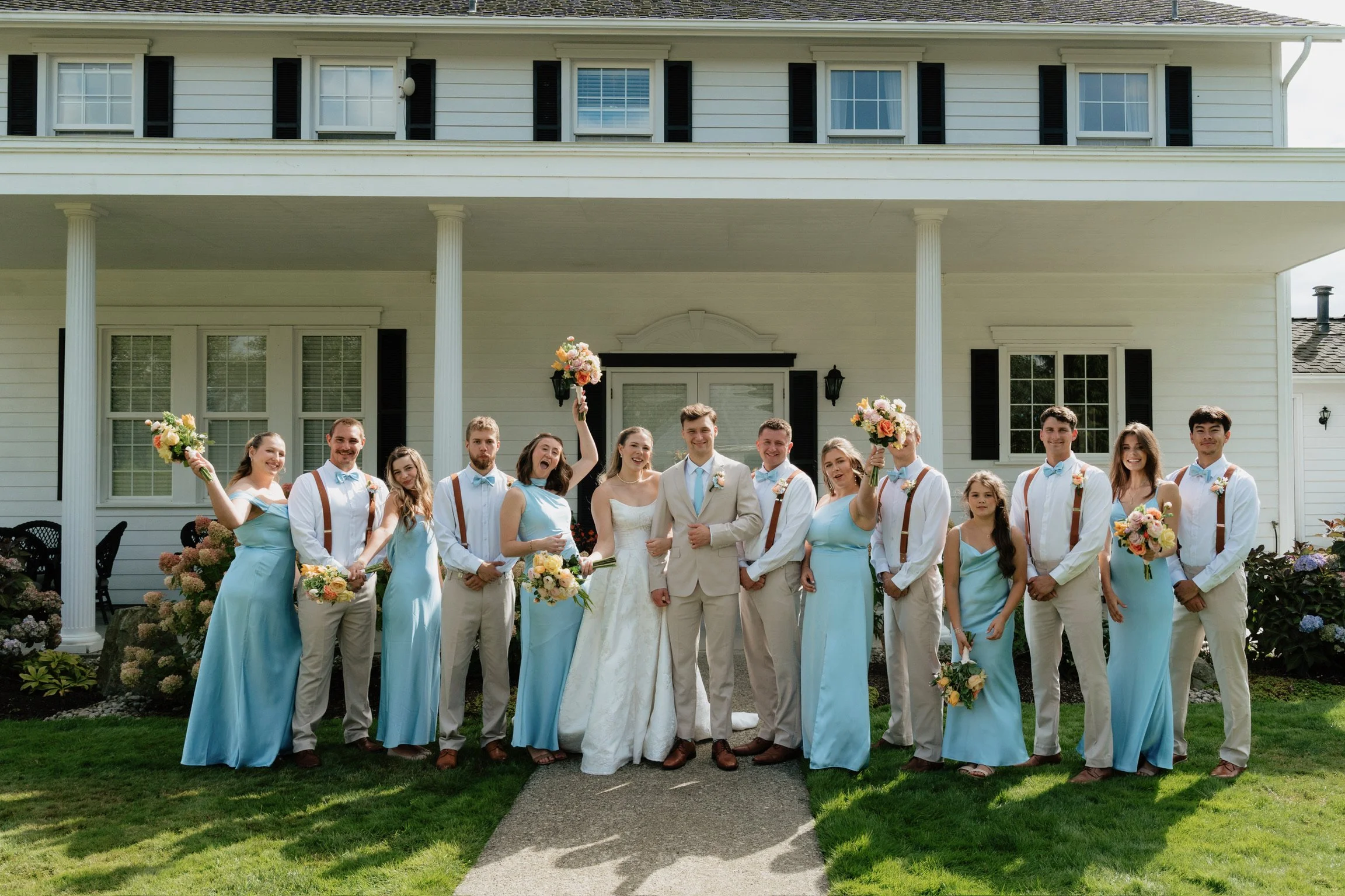A group of wedding guests and the bride and groom standing in front of a white house with columns, holding bouquets, smiling, and celebrating outdoors.