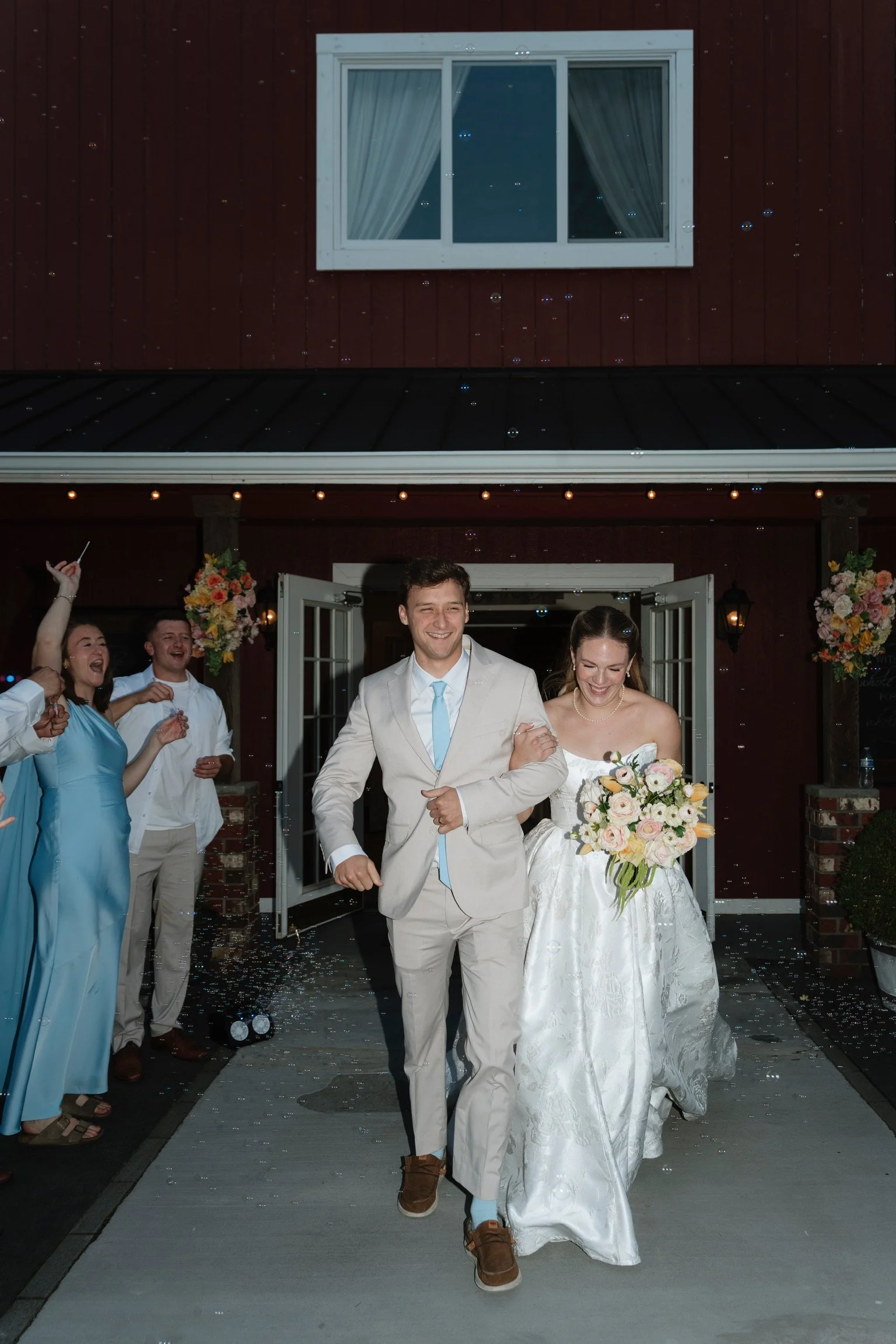 A newlywed couple is walking arm-in-arm out of a building at night, smiling, with guests celebrating around them and blowing bubbles. The bride is holding a bouquet of flowers, and the groom is wearing a light-colored suit with a blue tie. The buildi