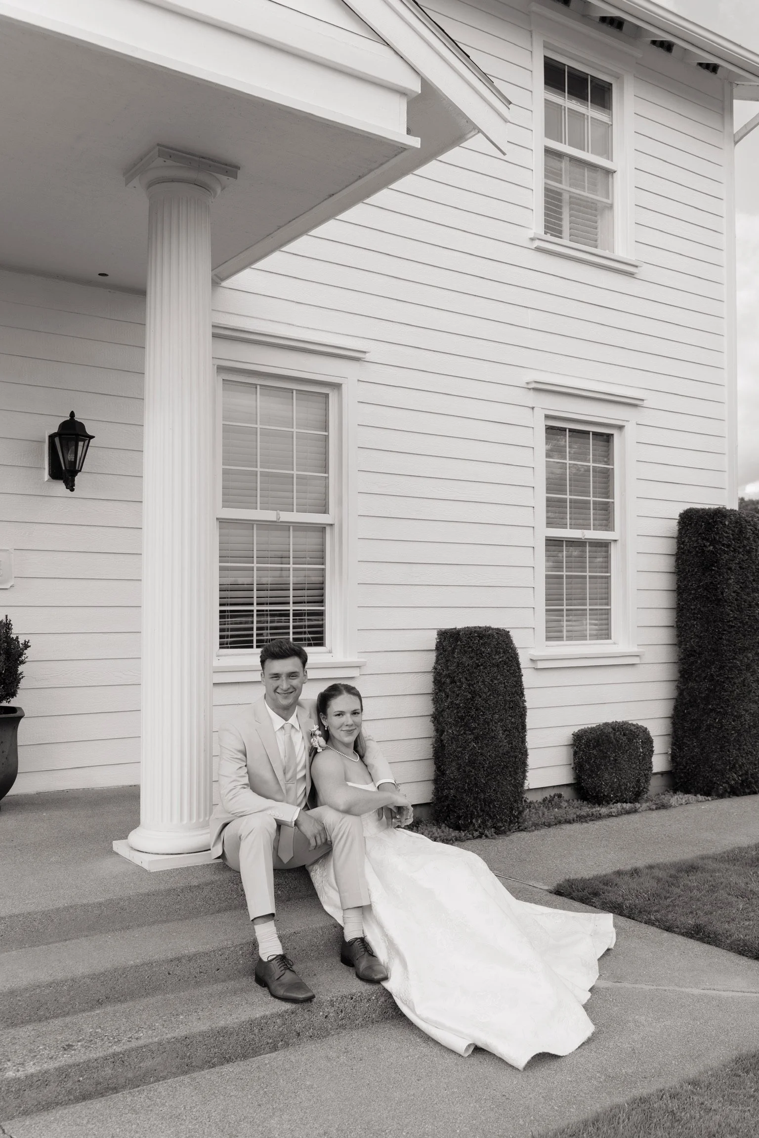 A happy bride and groom sitting on the sidewalk in front of a white house with green bushes. The groom is wearing a light-colored suit and the bride a white wedding dress.
