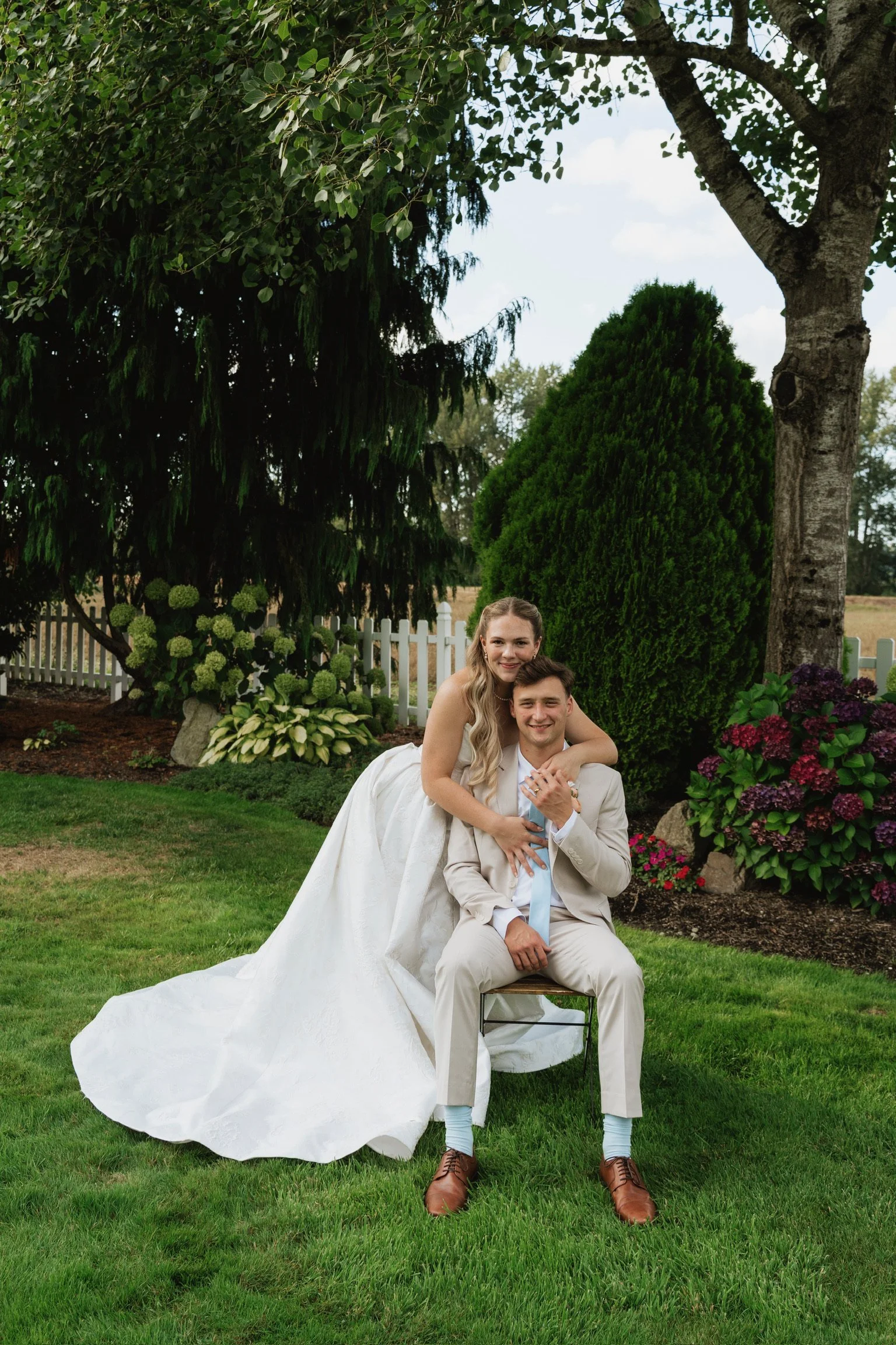 A newly married couple, bride and groom, posing together outdoors in a garden with trees, flowering bushes, a white picket fence, and a grassy lawn during daytime. The bride, wearing a white wedding gown, is sitting on the groom's lap who is seated o