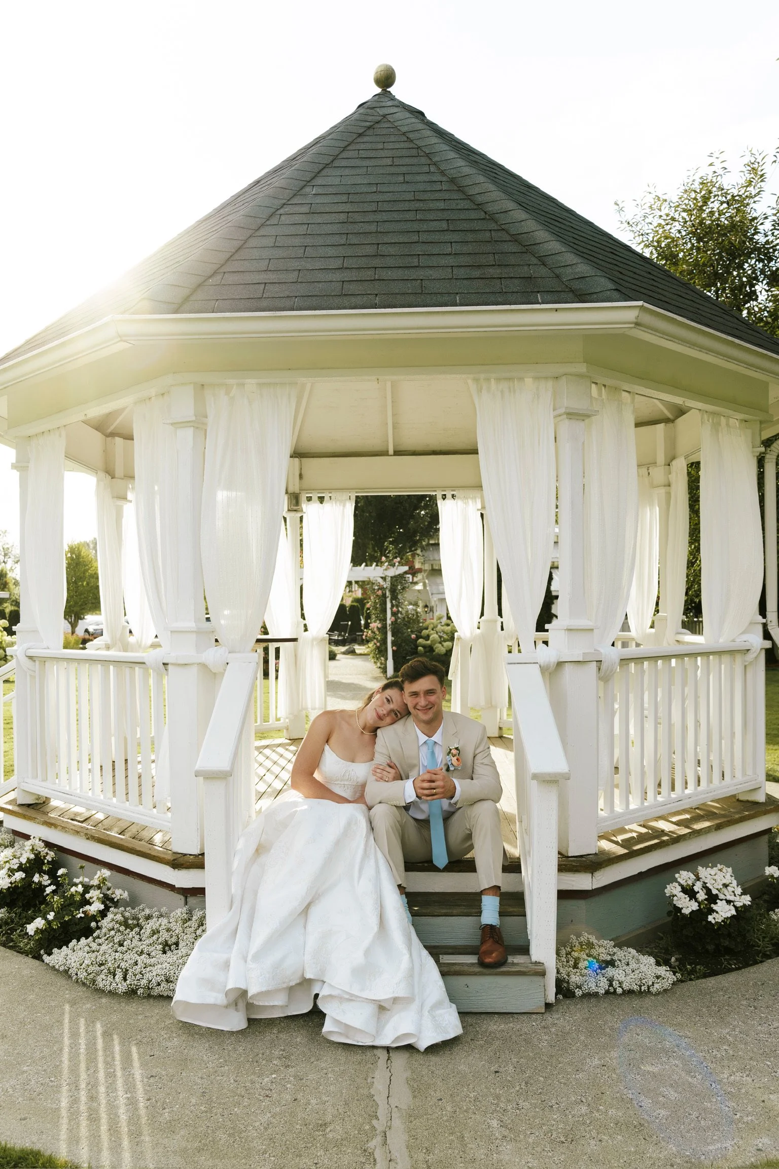 A newlywed couple sitting inside a white gazebo, with the bride leaning her head on the groom's shoulder, both smiling, during their wedding photo shoot on a sunny day.