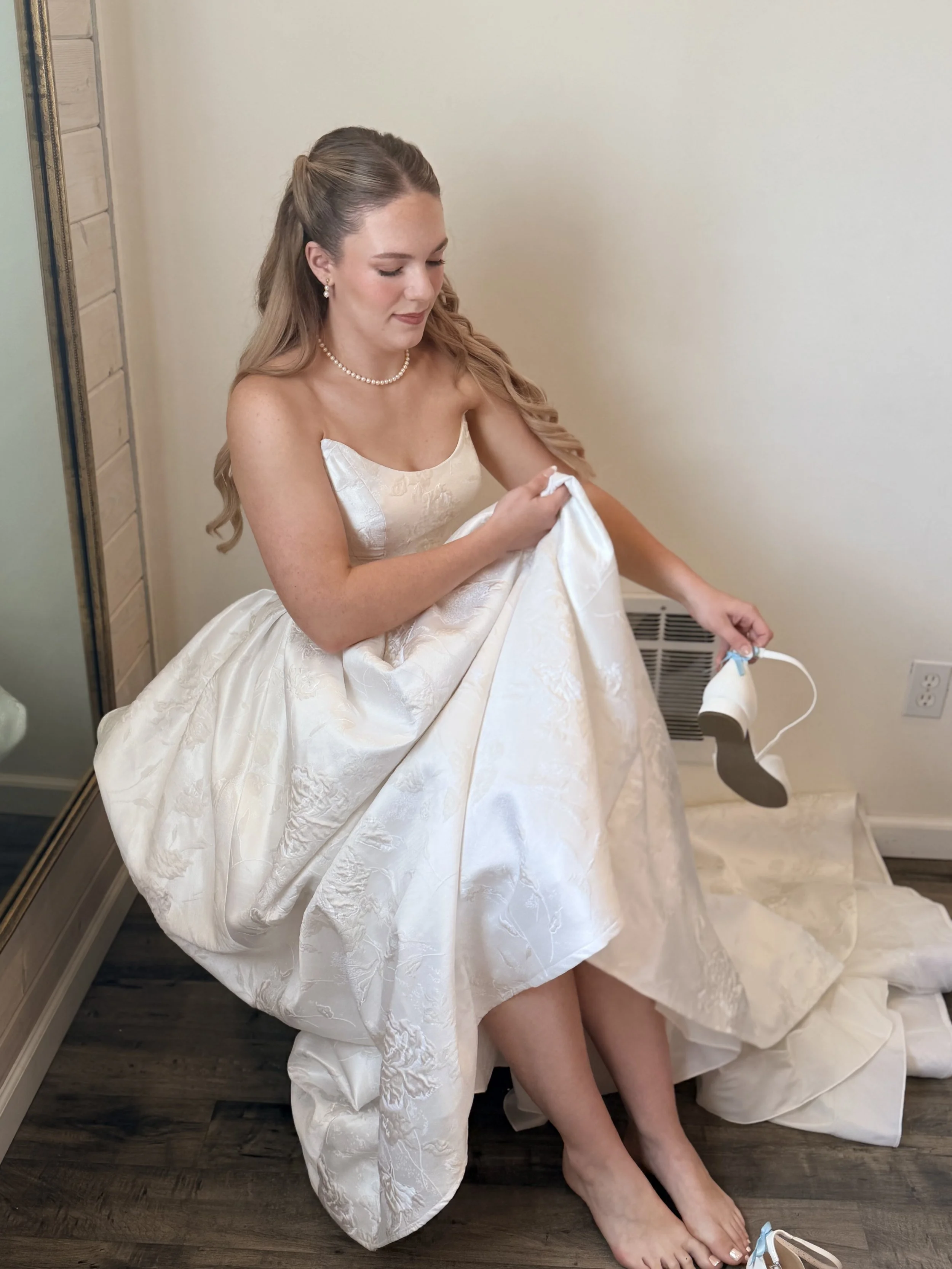 A woman in a white wedding dress is sitting on the floor, adjusting her dress, with her legs slightly bent. She has long wavy hair and is wearing pearl earrings and a pearl necklace. There is a white shoe with blue ribbon on the floor near her foot, 