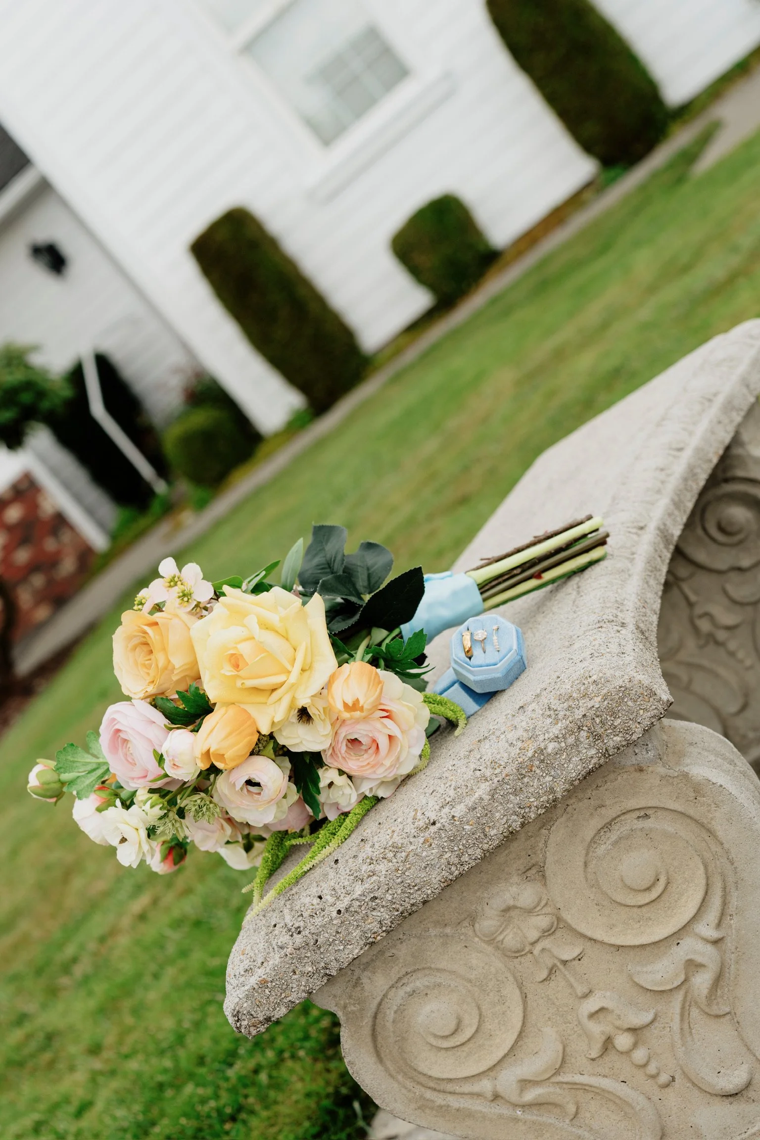 A bouquet of yellow, pink, and white flowers placed on a carved stone bench in a garden with trimmed bushes and a white house in the background.