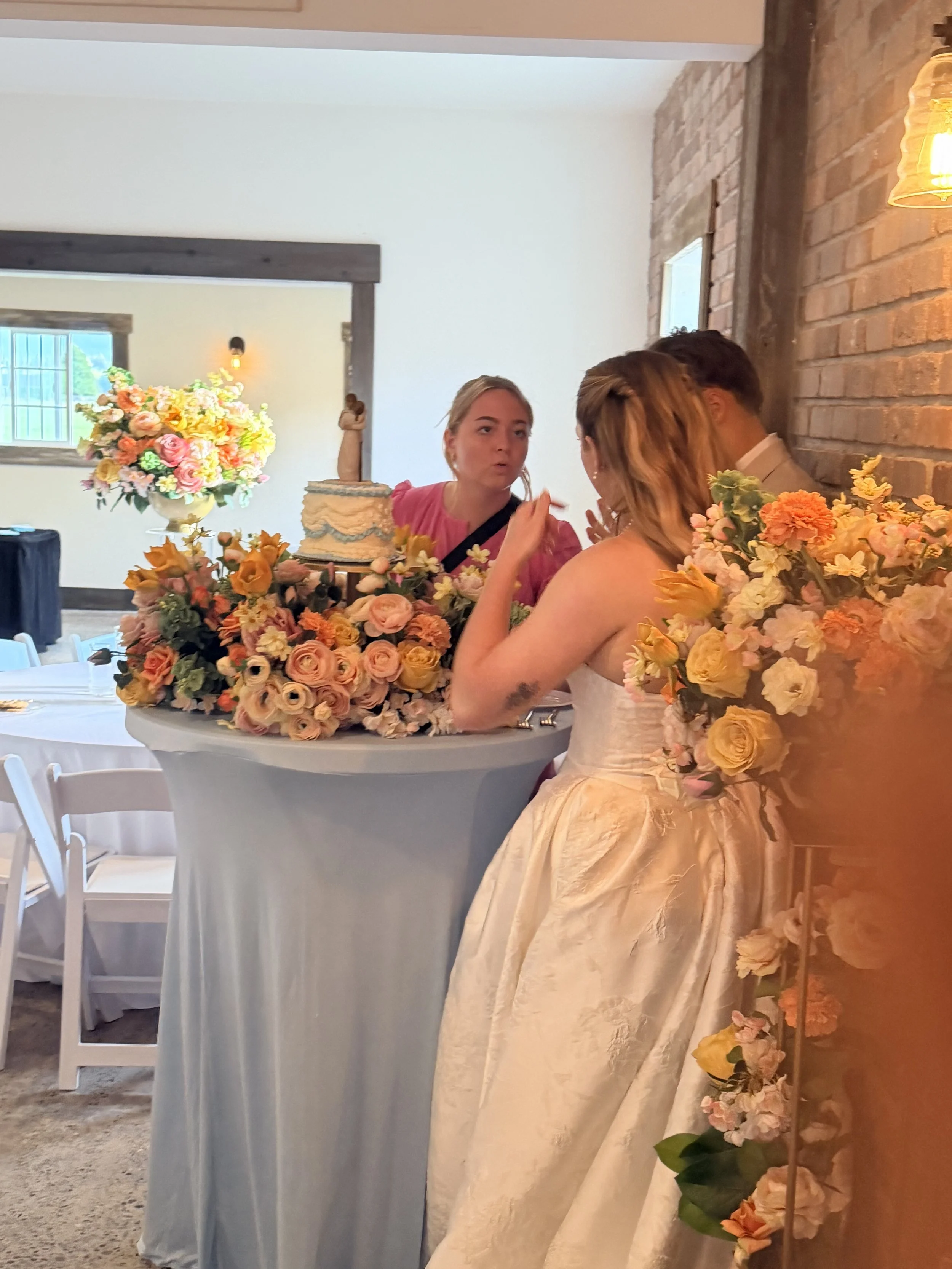A wedding reception scene showing a woman in a white bridal gown with a floral bouquet, a lady in pink, and a man in a light-colored suit, with a decorated table and floral arrangements.
