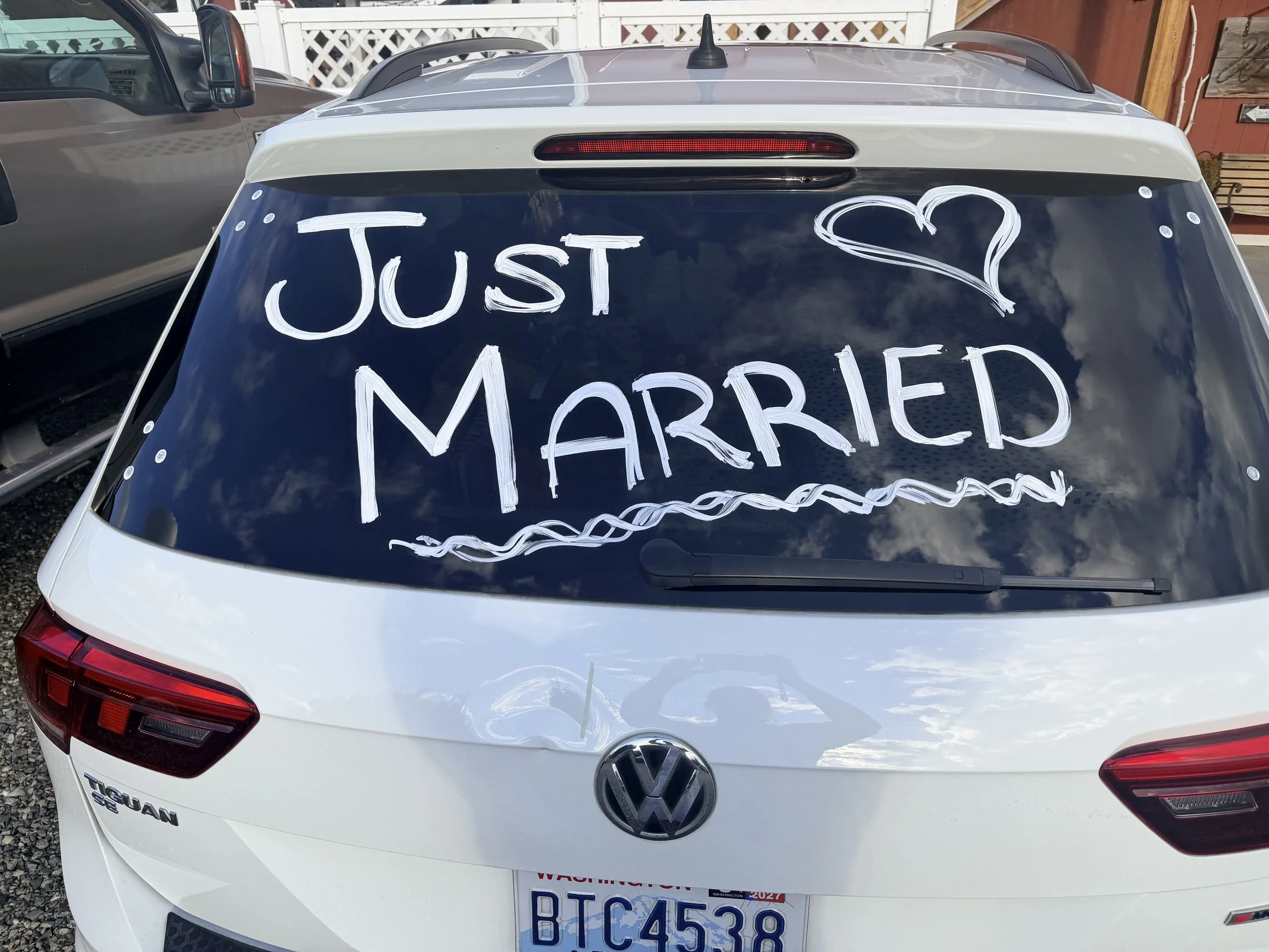 The rear of a white Volkswagen Tiguan SUV with a sign written in white marker that says, "Just ♥ Married," on the back windshield.