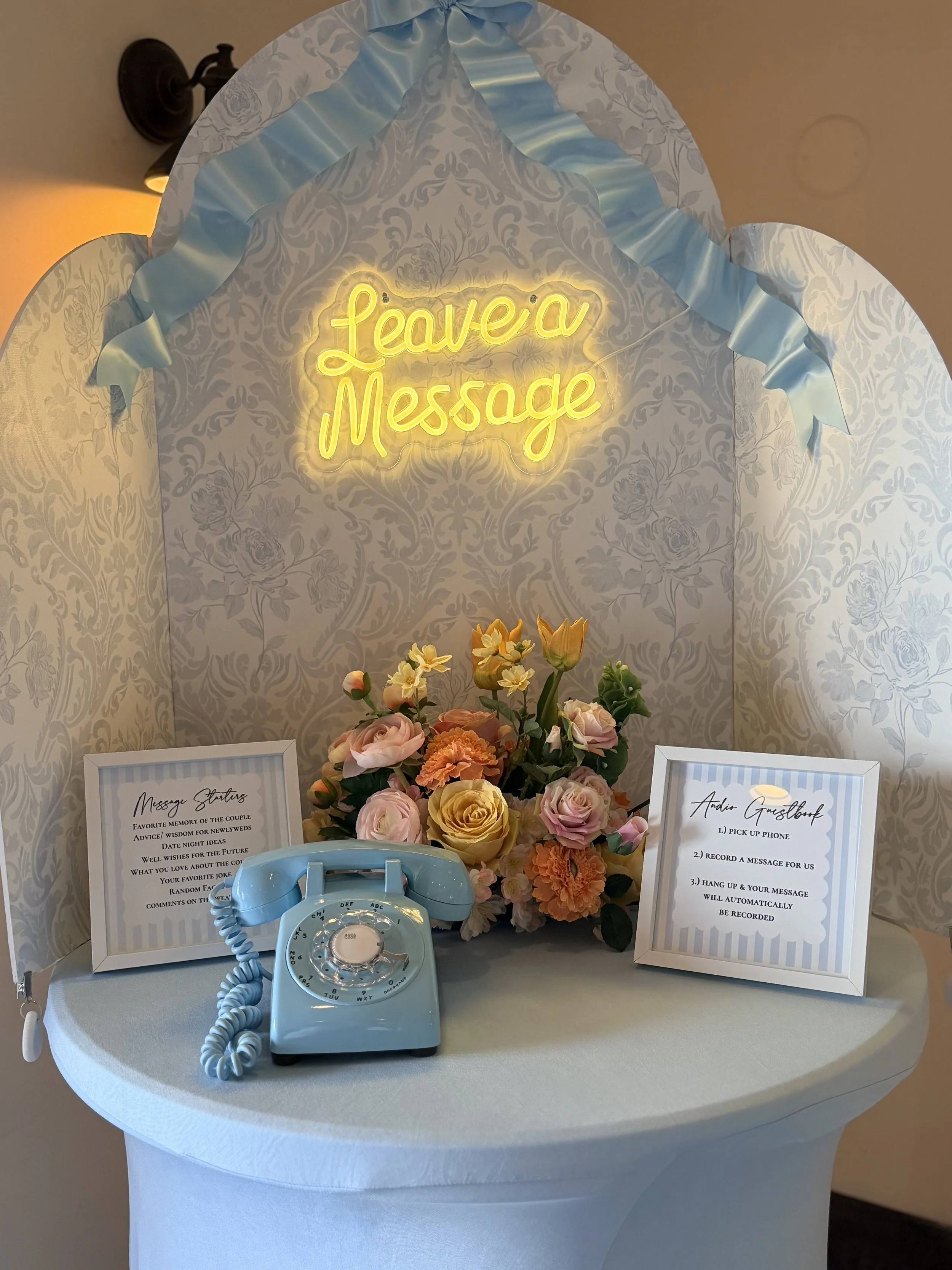 A vintage turquoise rotary telephone resting on a white table, with framed signs and a large floral centerpiece behind it, and a neon sign that reads 'Leave a Message' on a patterned backdrop.