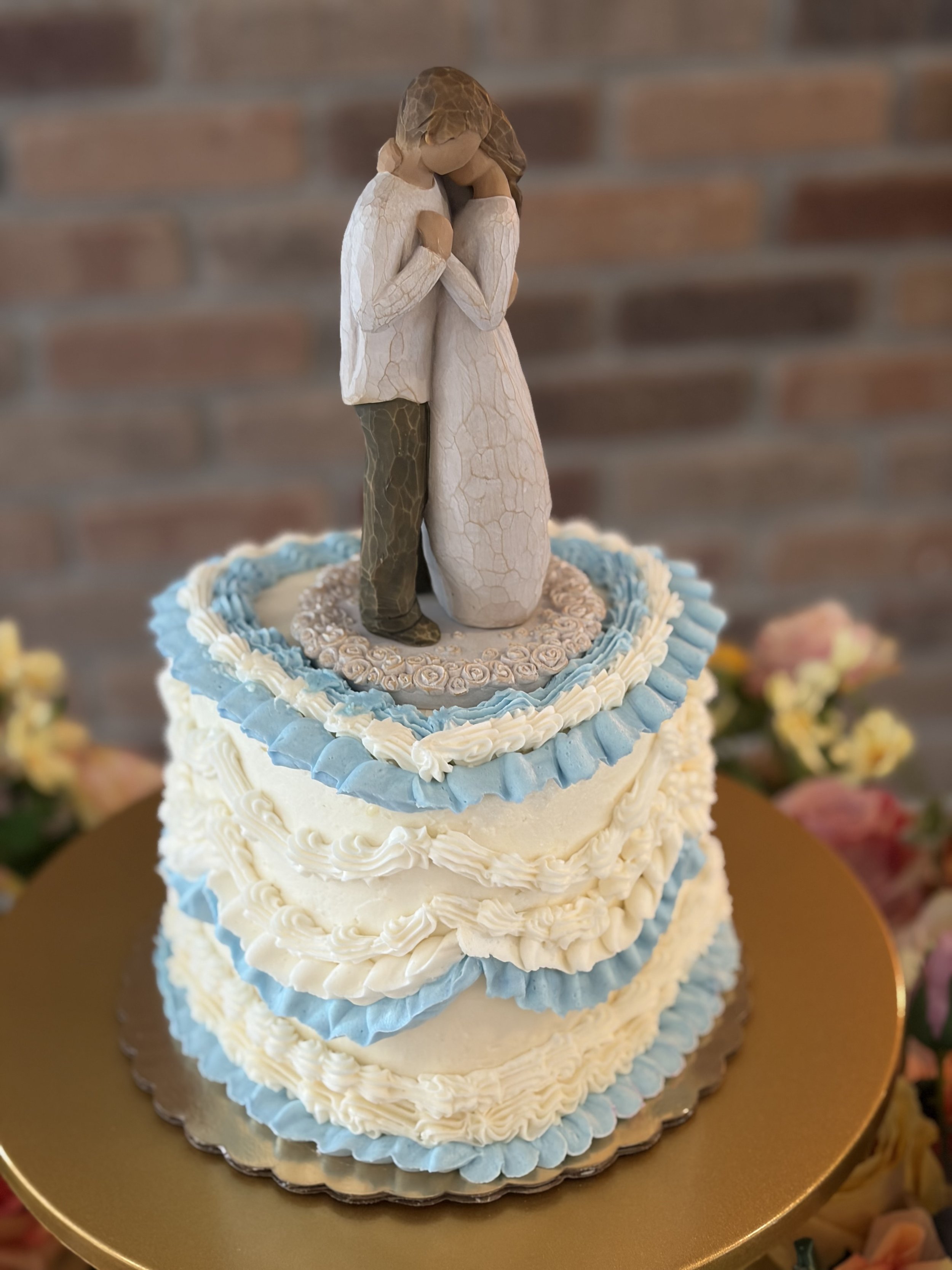 A wedding cake with a cake topper of a couple hugging, on a gold cake board, with white and blue decorative frosting.