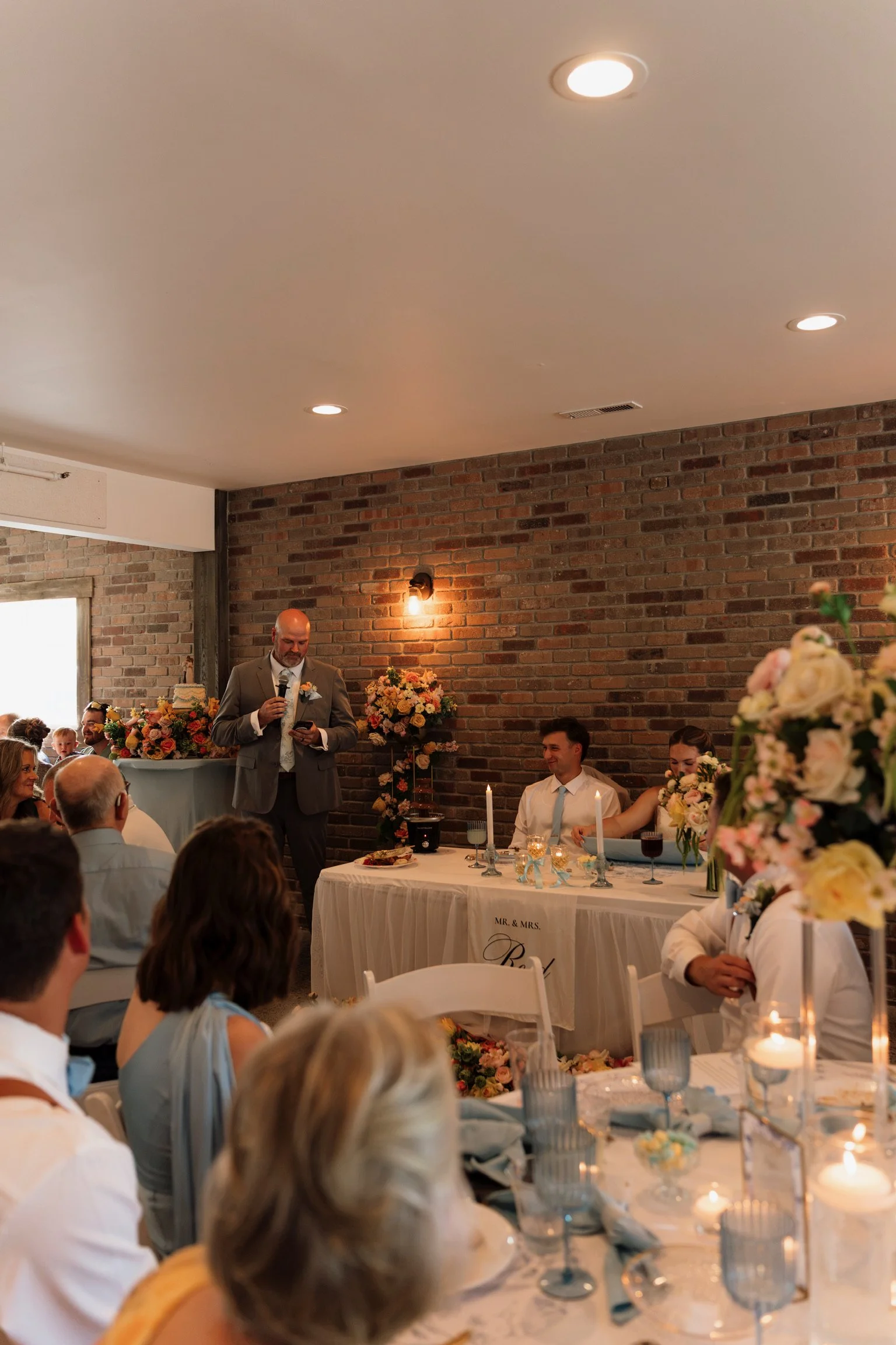 A wedding reception in a rustic venue with exposed brick walls, featuring a long table decorated with flowers and candles, and a man in a suit giving a speech.