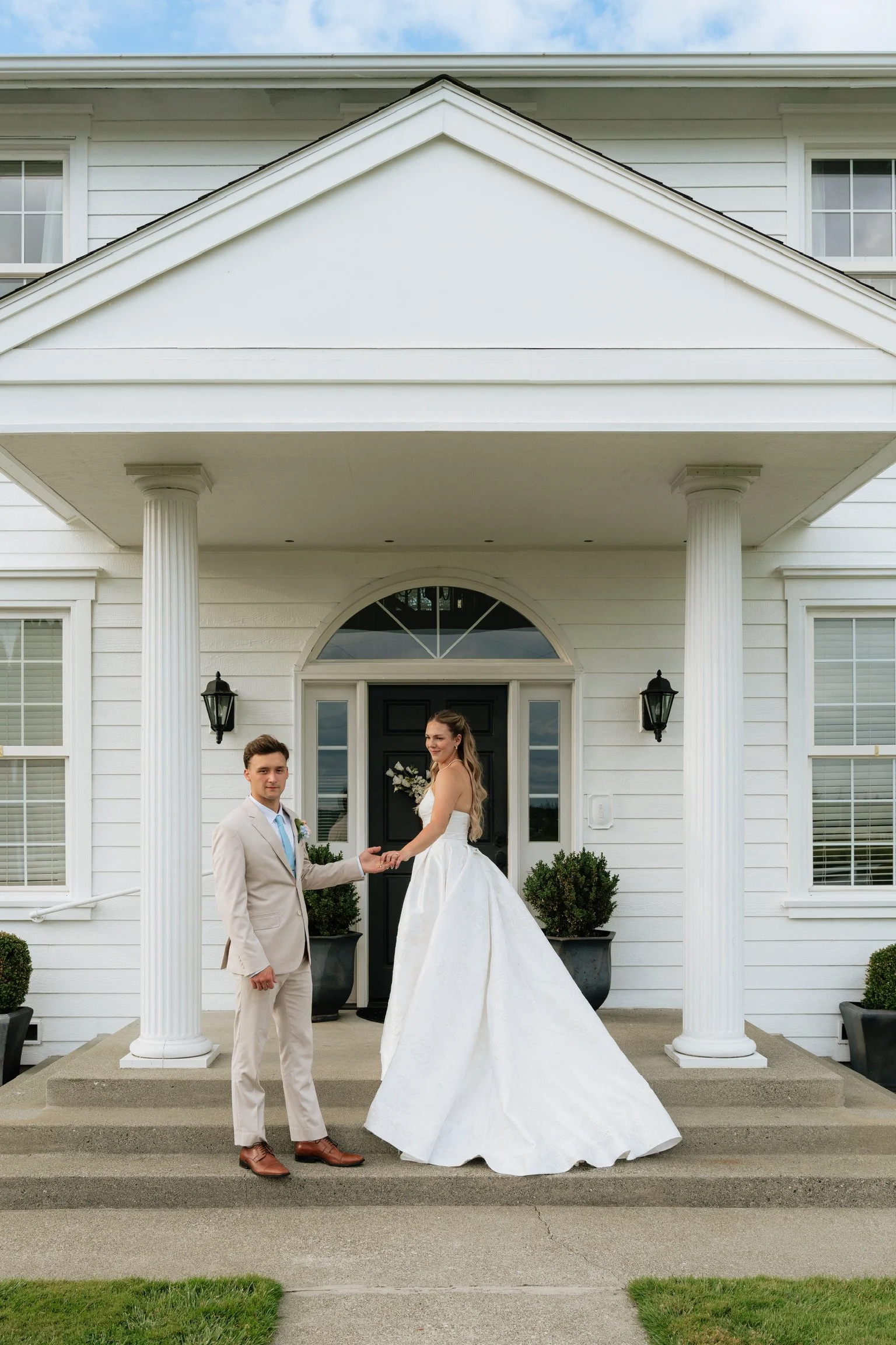 A couple dressed in wedding attire standing in front of a white house with columns, holding hands and smiling, with the bride in a white gown and the groom in a light-colored suit.