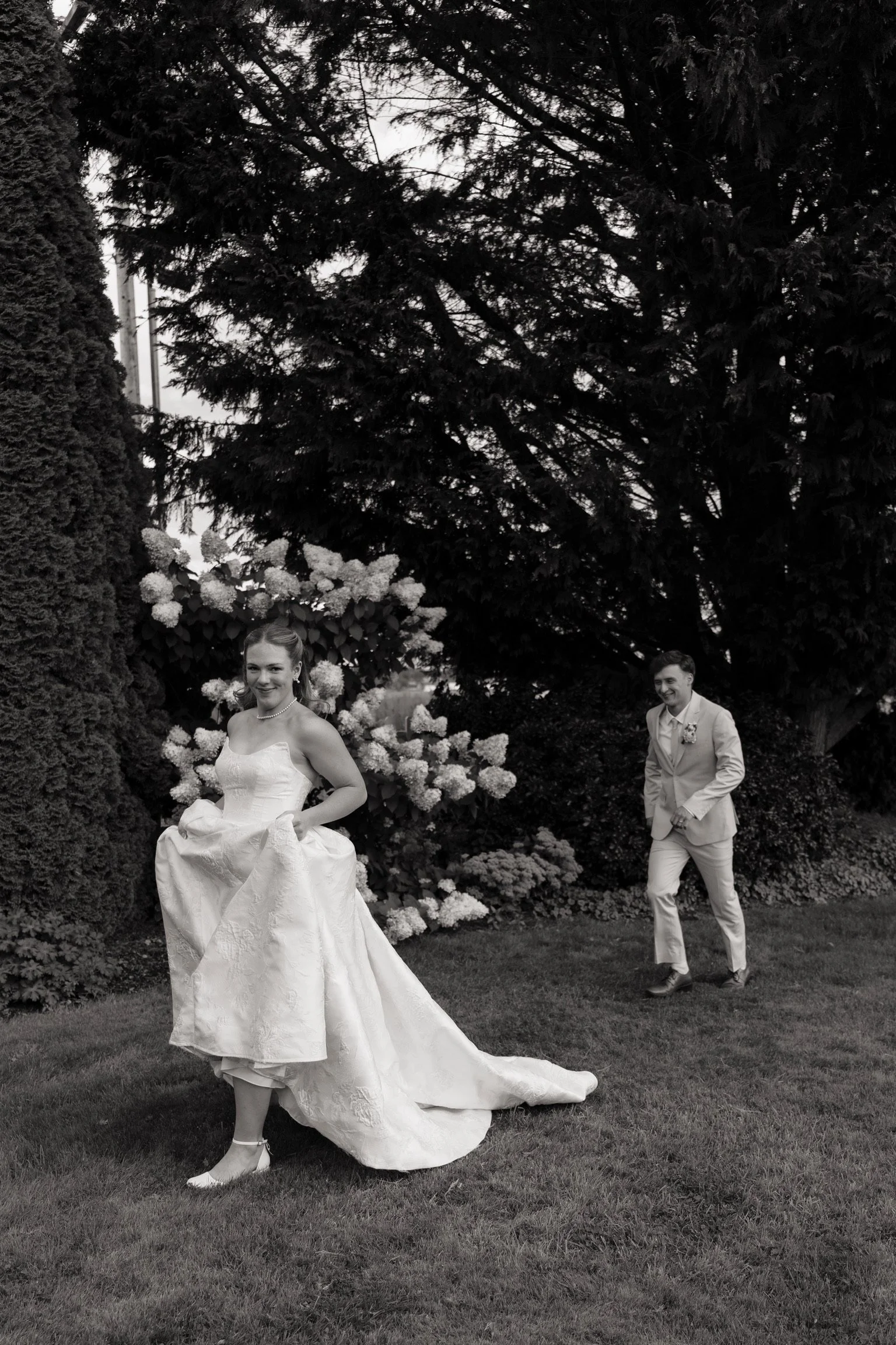 A bride in a white wedding gown and a groom in a light-colored suit walking outdoors in a garden with lush greenery and large trees in the background.