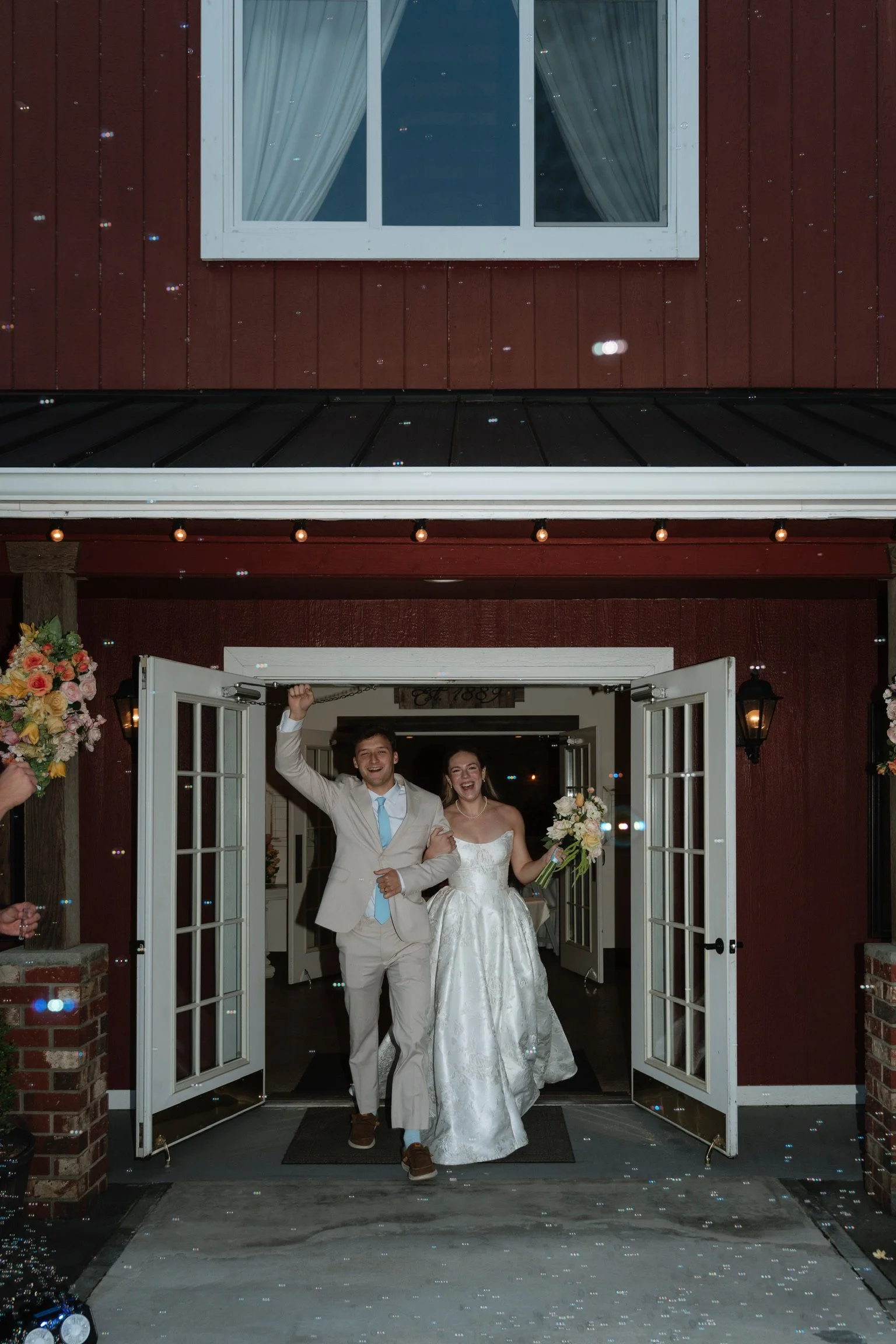 A newlywed couple, a man in a light-colored suit and a woman in a strapless wedding dress, walking out of a building with open double doors, smiling and celebrating.