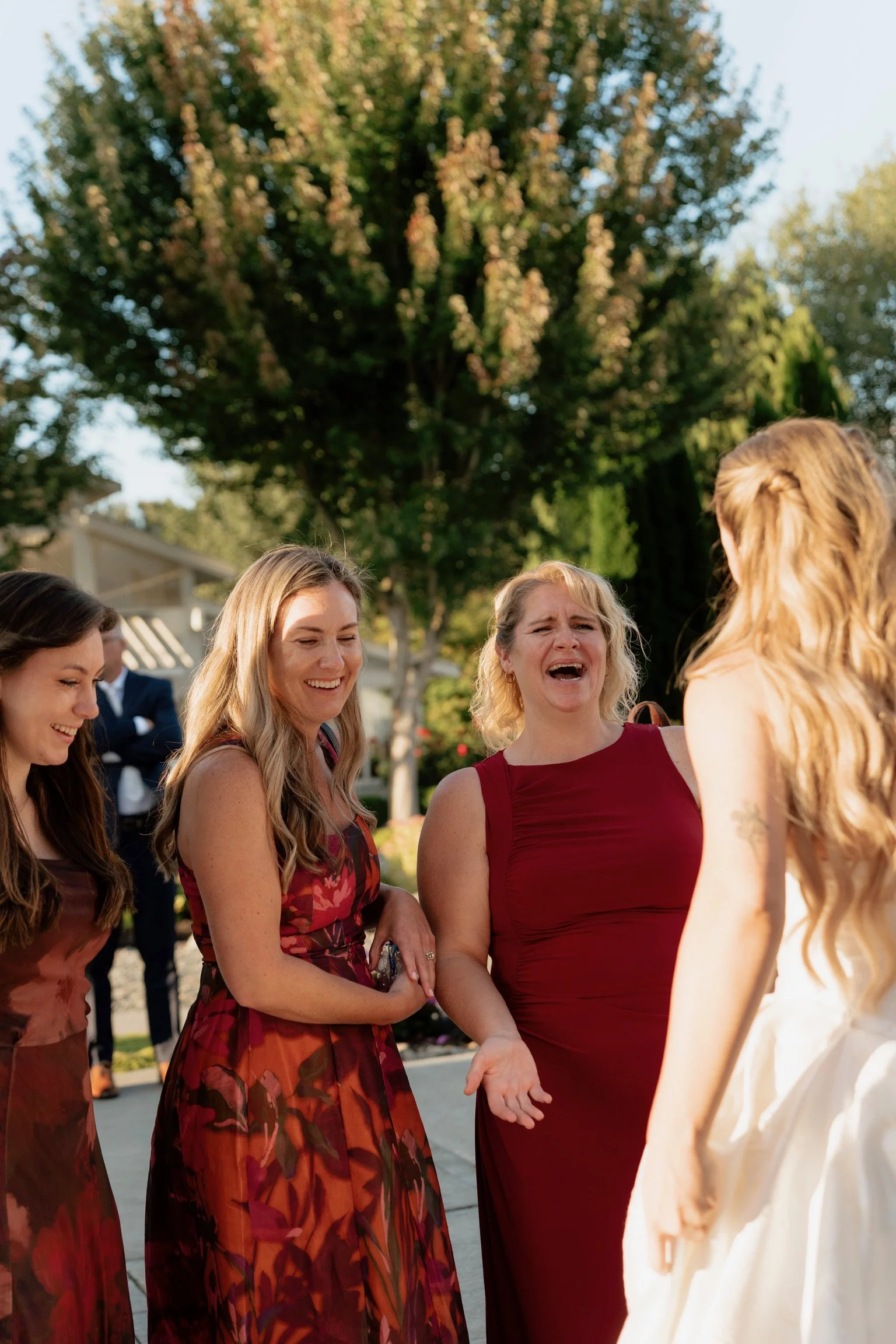 A group of women dressed in formal attire, smiling and laughing, engaging in conversation outdoors under a large tree during daytime.