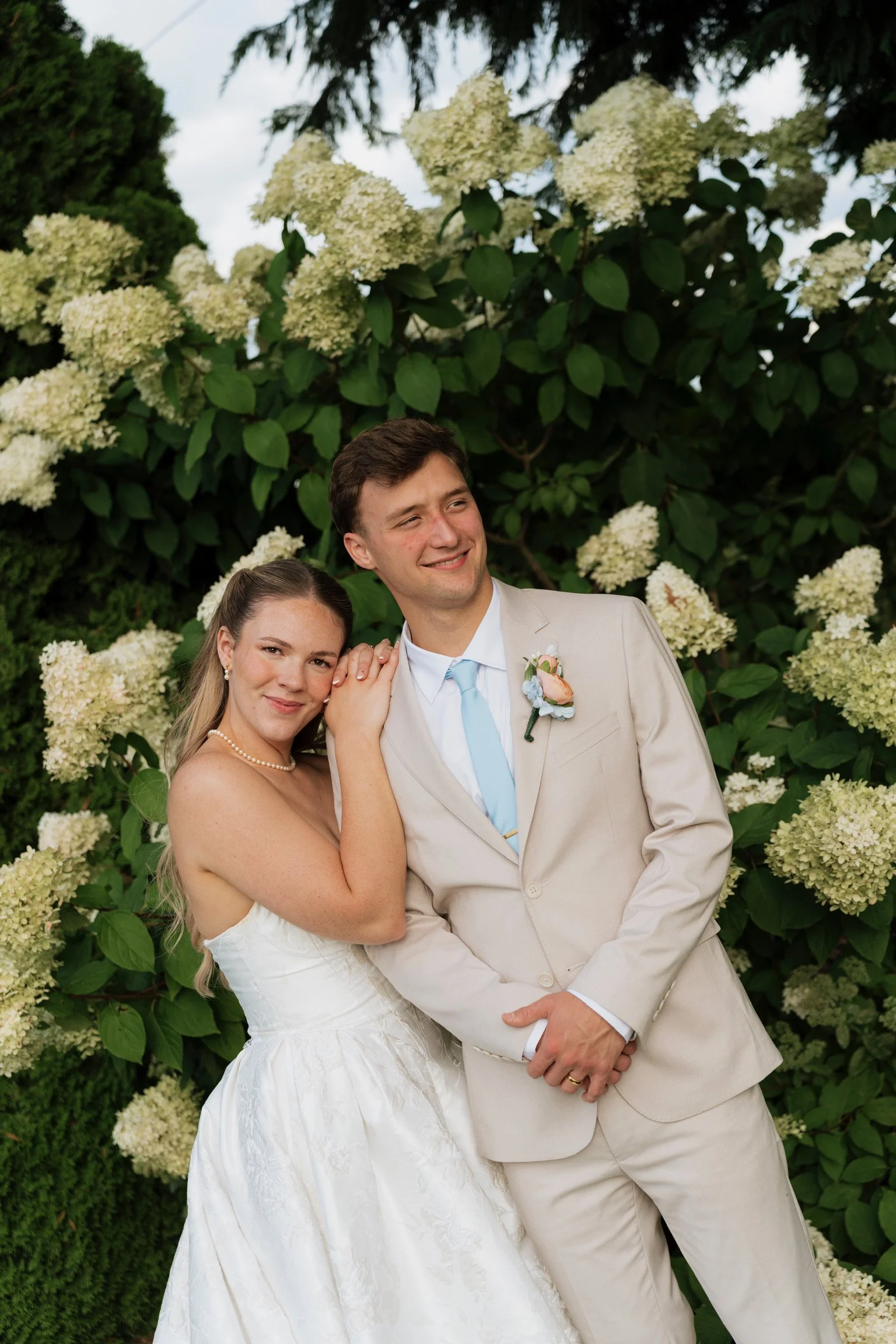 A newlywed couple smiling and posing in front of a lush bush with white flowers; the bride is in a white wedding dress with a pearl necklace, and the groom is in a beige suit with a light blue tie and boutonniere.
