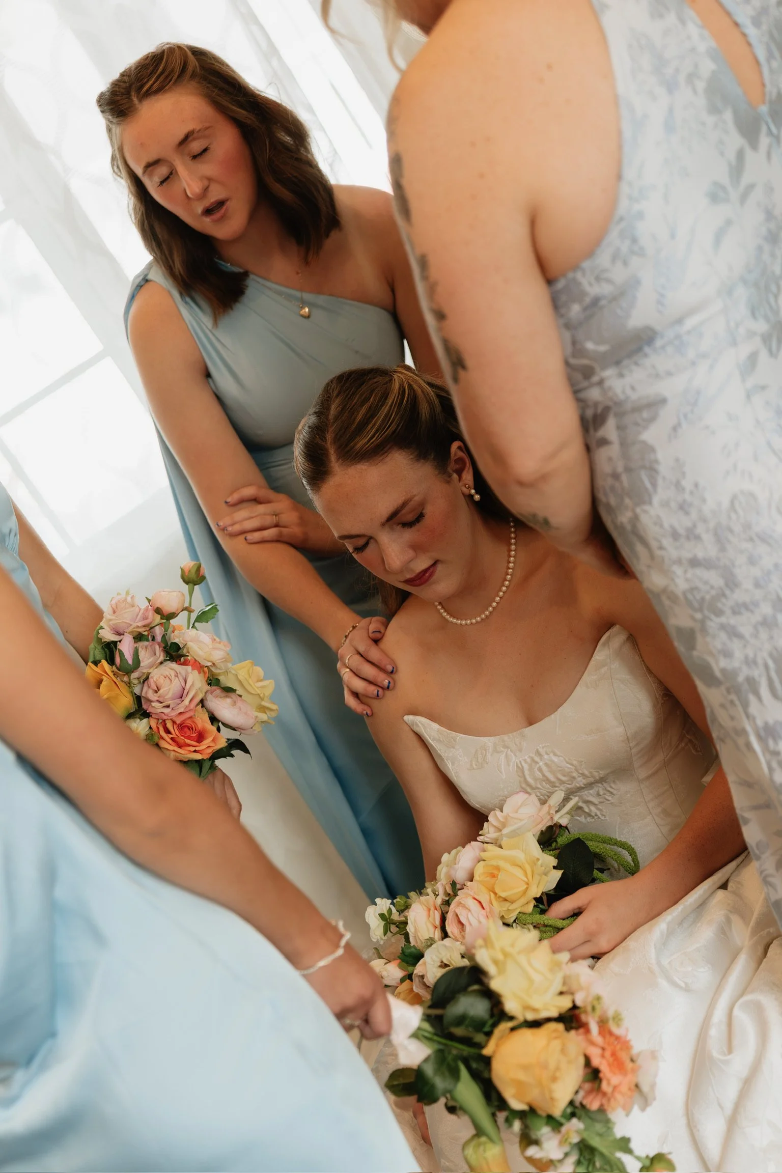 A bride dressed in a white wedding gown holding a bouquet of pink, peach, and cream roses, surrounded by women in pastel dresses during a wedding ceremony.