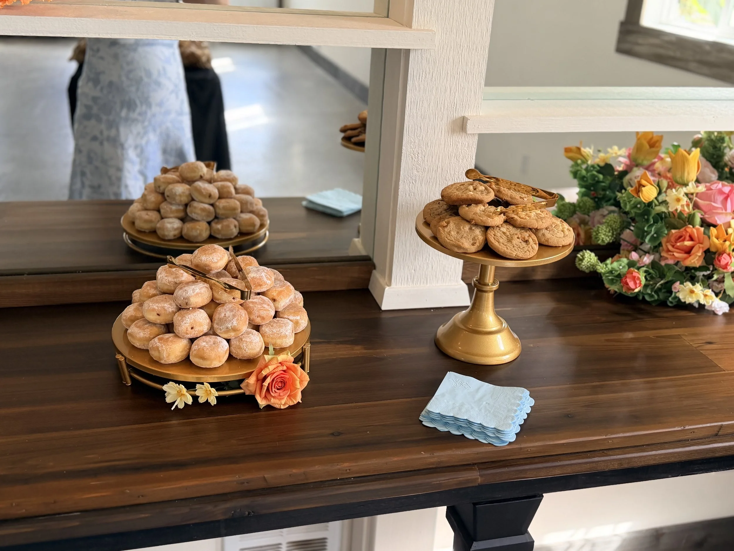 Assortment of cookies on gold cake stands, decorated with flowers, on a wooden table near a mirror.