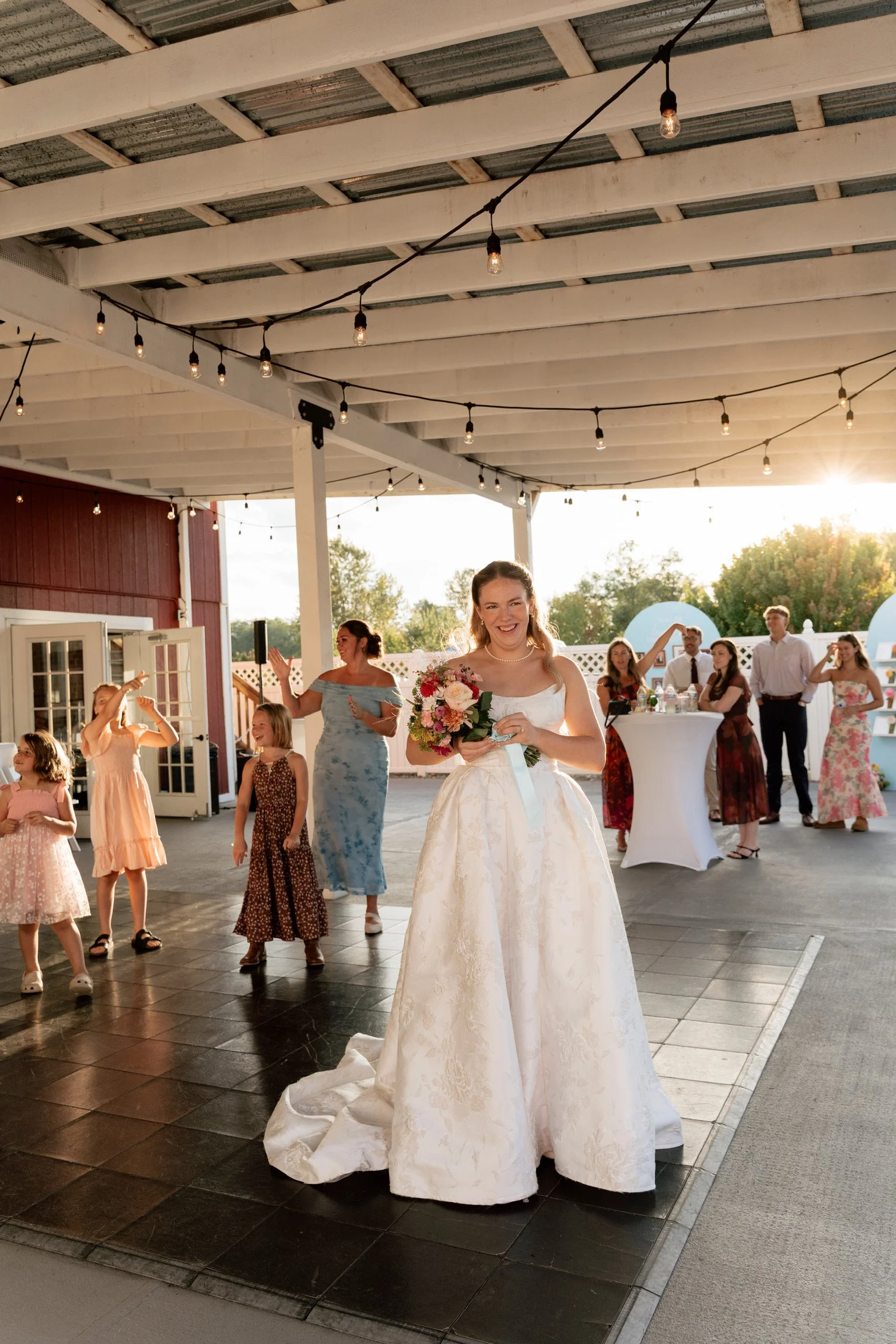 A bride in a white wedding dress holding a bouquet of flowers at an outdoor wedding reception, with guests dancing and socializing under a covered patio with string lights and a rustic setting.
