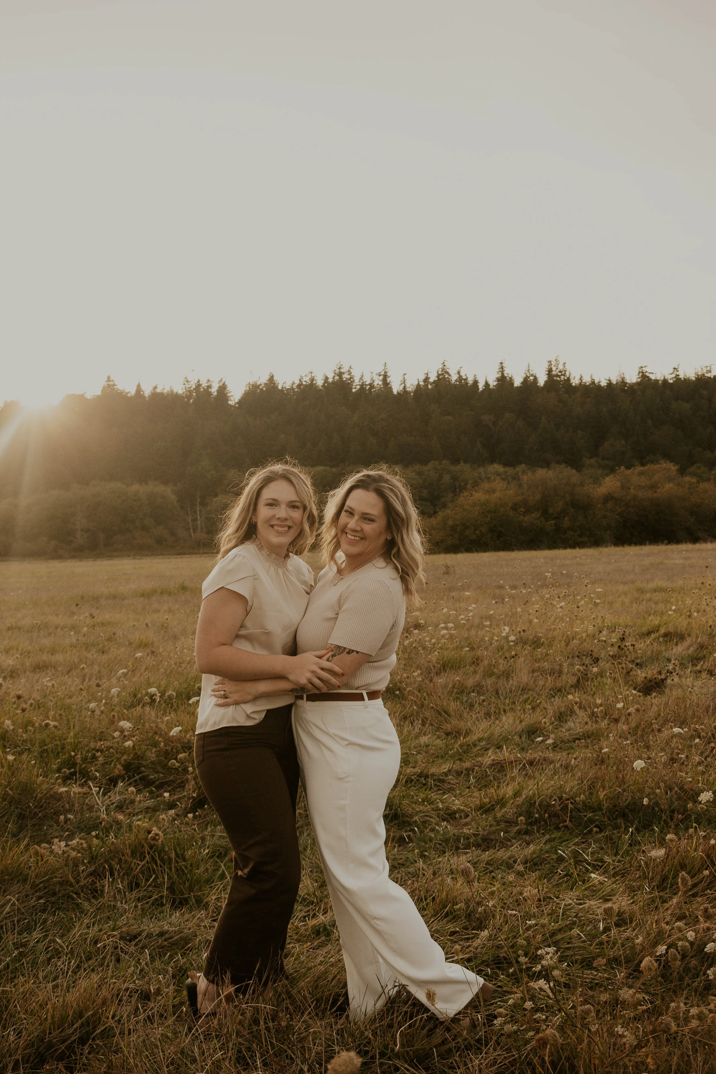 Two women smiling and hugging in a grassy field with wildflowers, during golden hour with the sun setting behind a forested hill in the background.